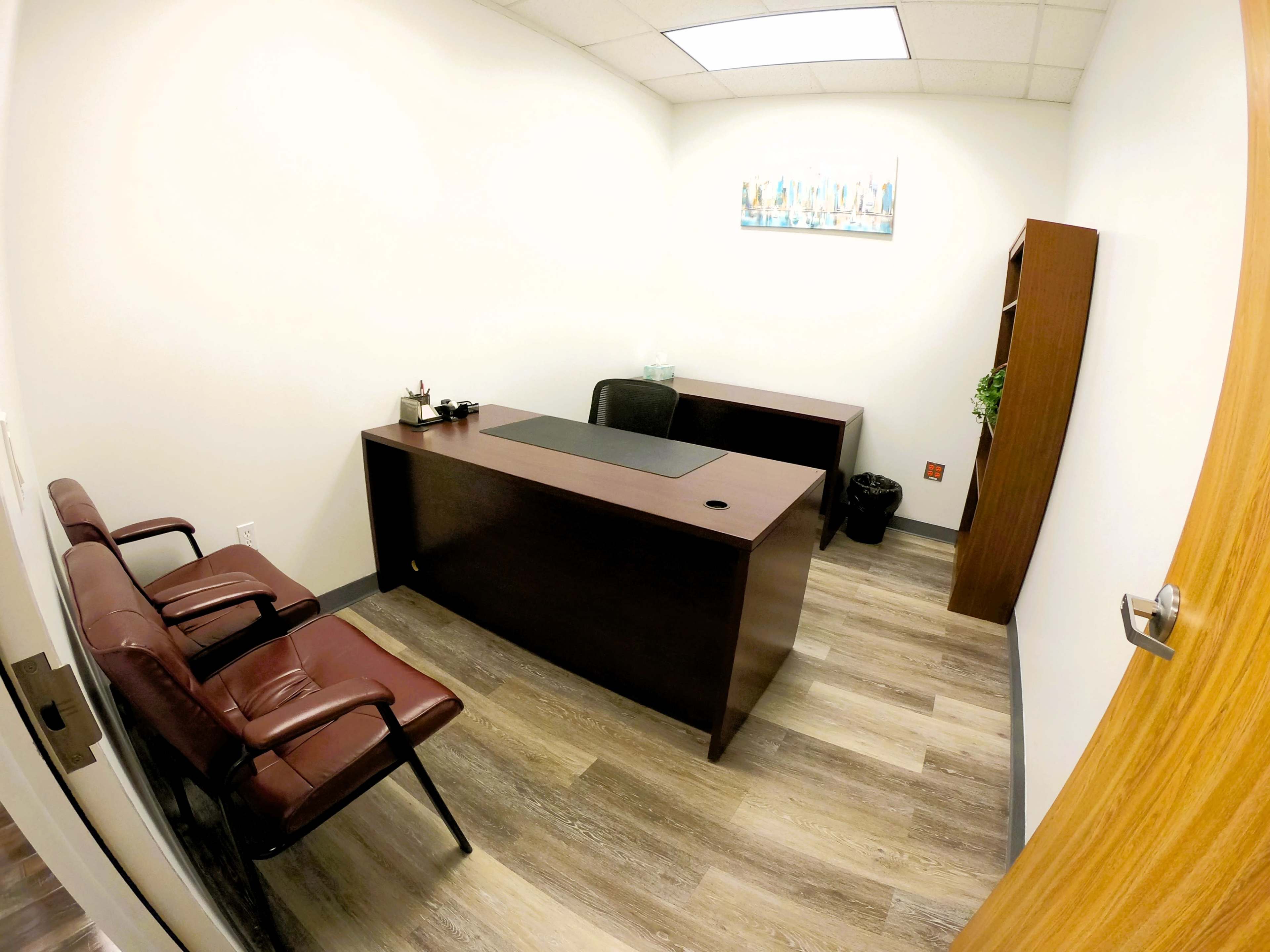 A small office with a large desk, two brown chairs, a bookcase, and a wall-mounted painting.