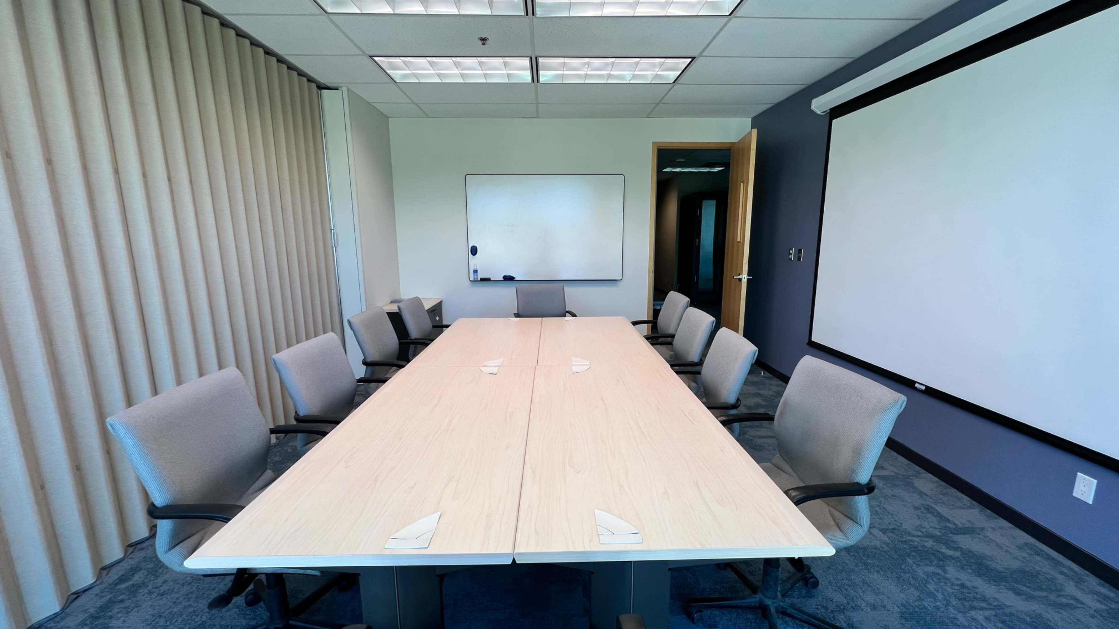 A conference room features a long wooden table surrounded by eight gray chairs, with a whiteboard and a large projection screen on adjacent walls.