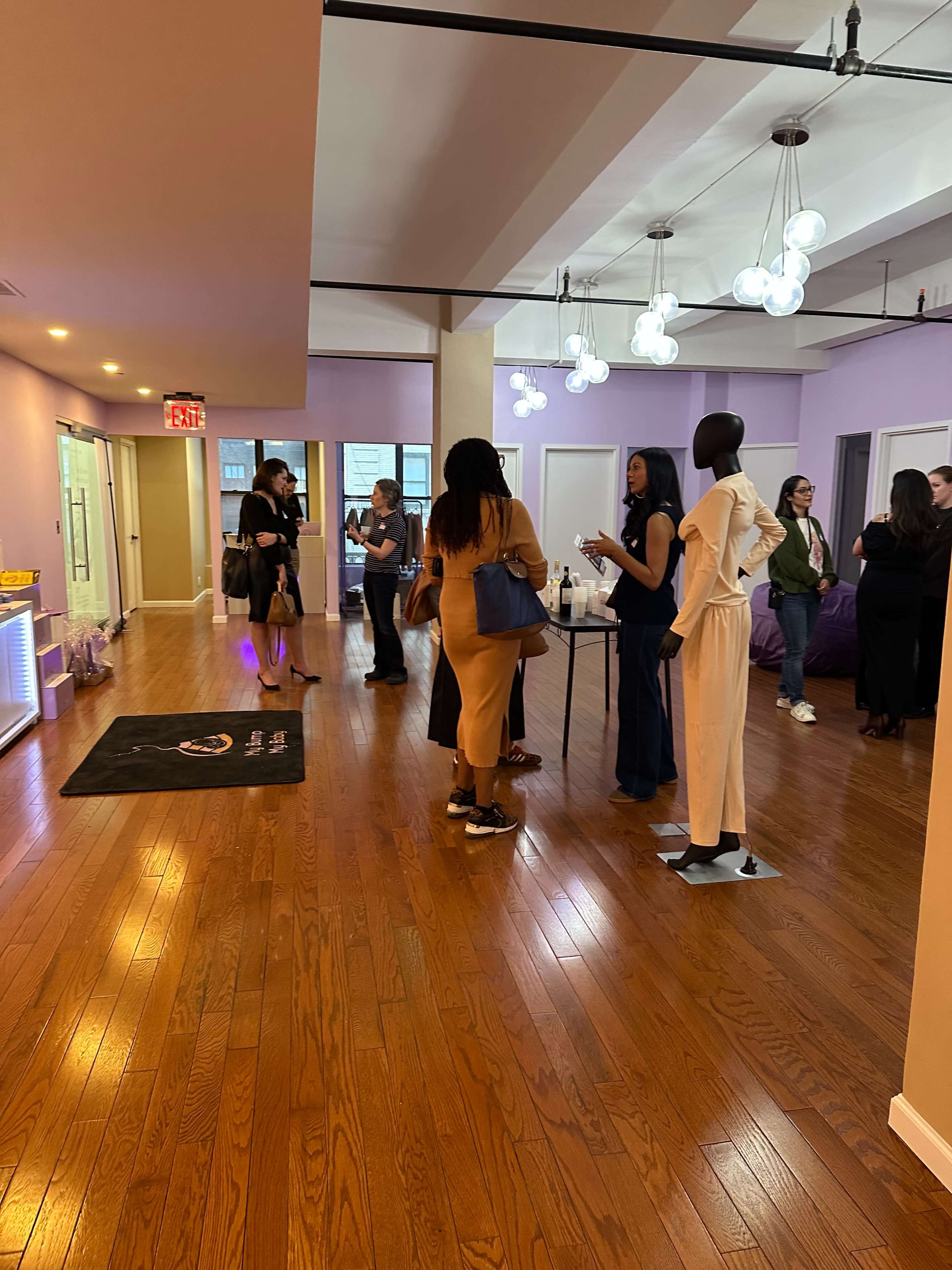 The image shows a modern, brightly lit interior space with people gathered around a table, socializing, and displaying a mannequin in stylish clothing.