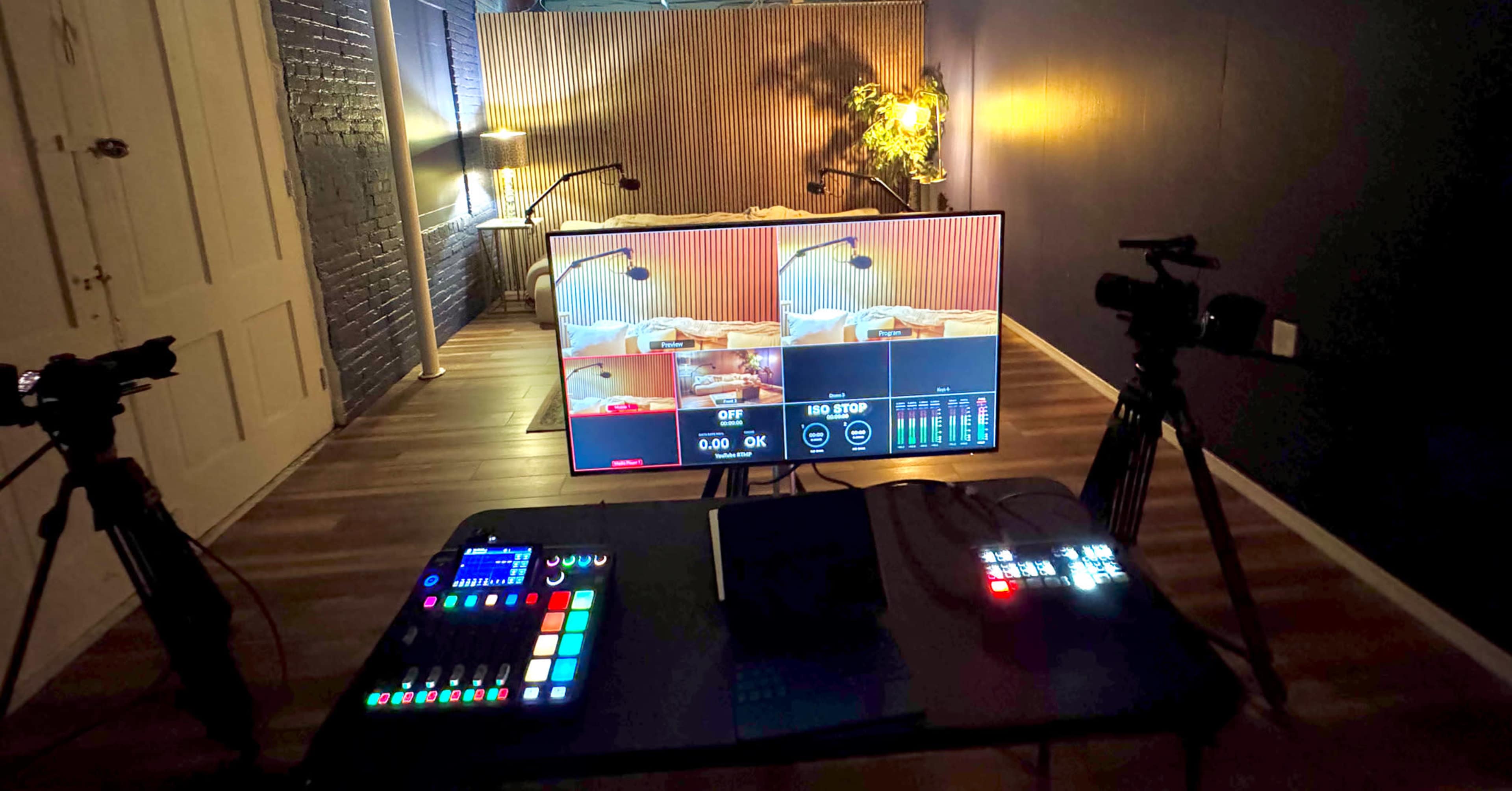 The image shows a video production setup with a control panel and two cameras facing a large screen displaying a live feed in a dimly lit room.