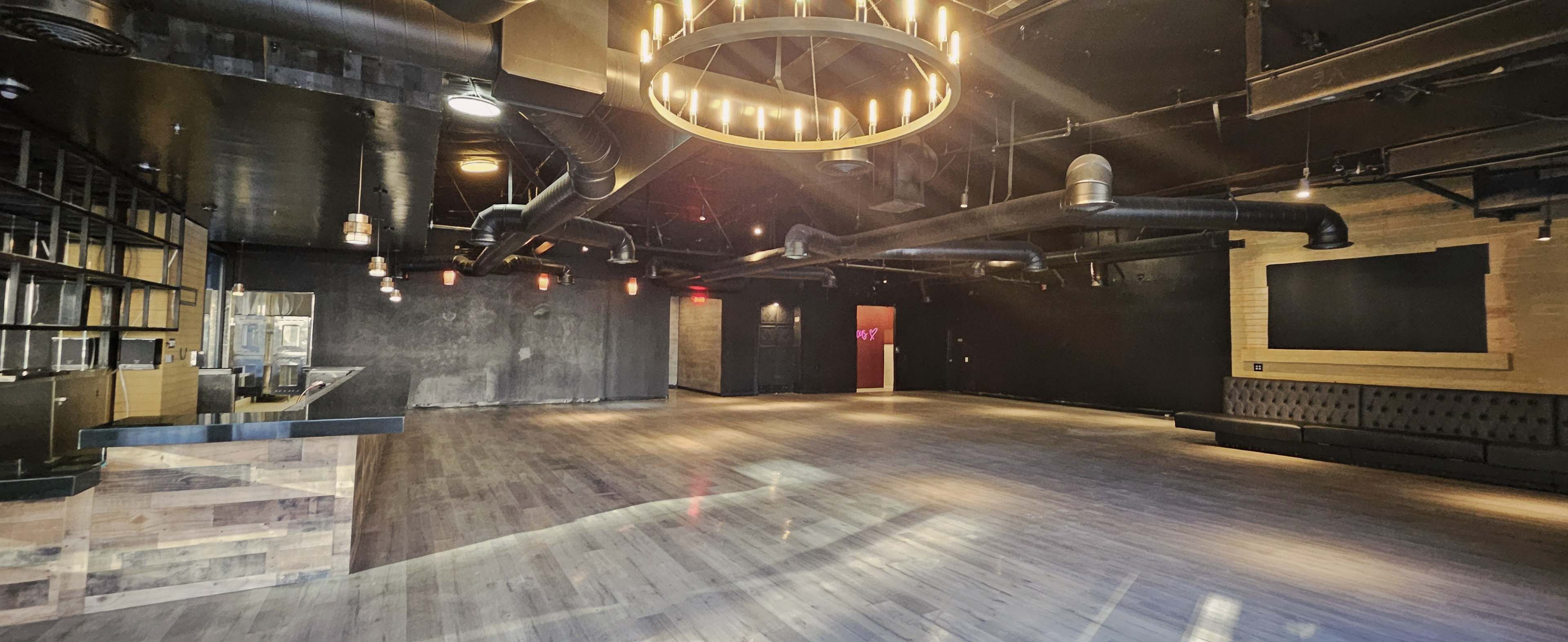 The image shows a spacious, dimly lit interior featuring a wooden floor, a circular chandelier, and a bar area against one wall.