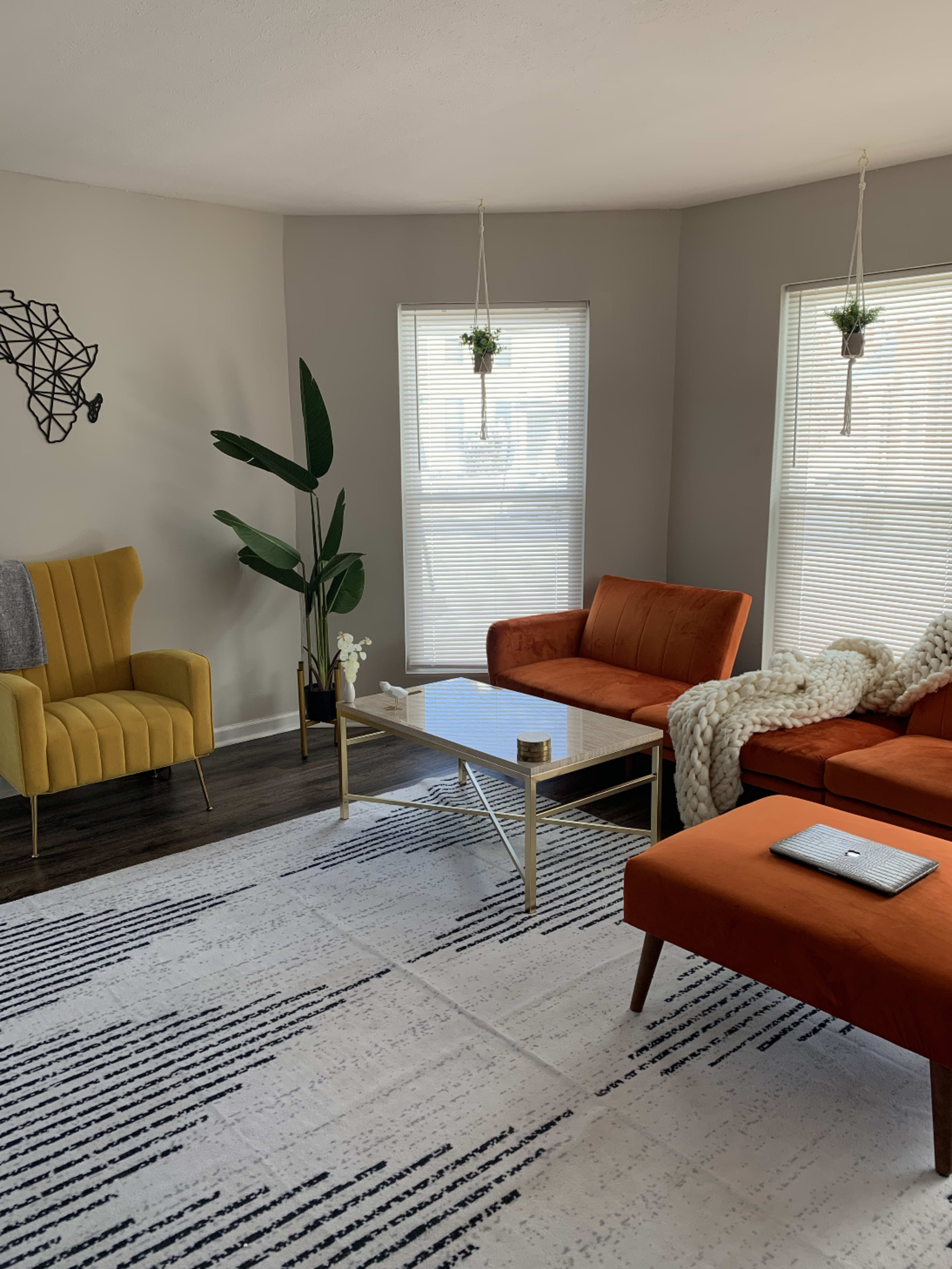 The living room features orange and yellow seating, a glass coffee table, a textured area rug, and large windows with subtle greenery.
