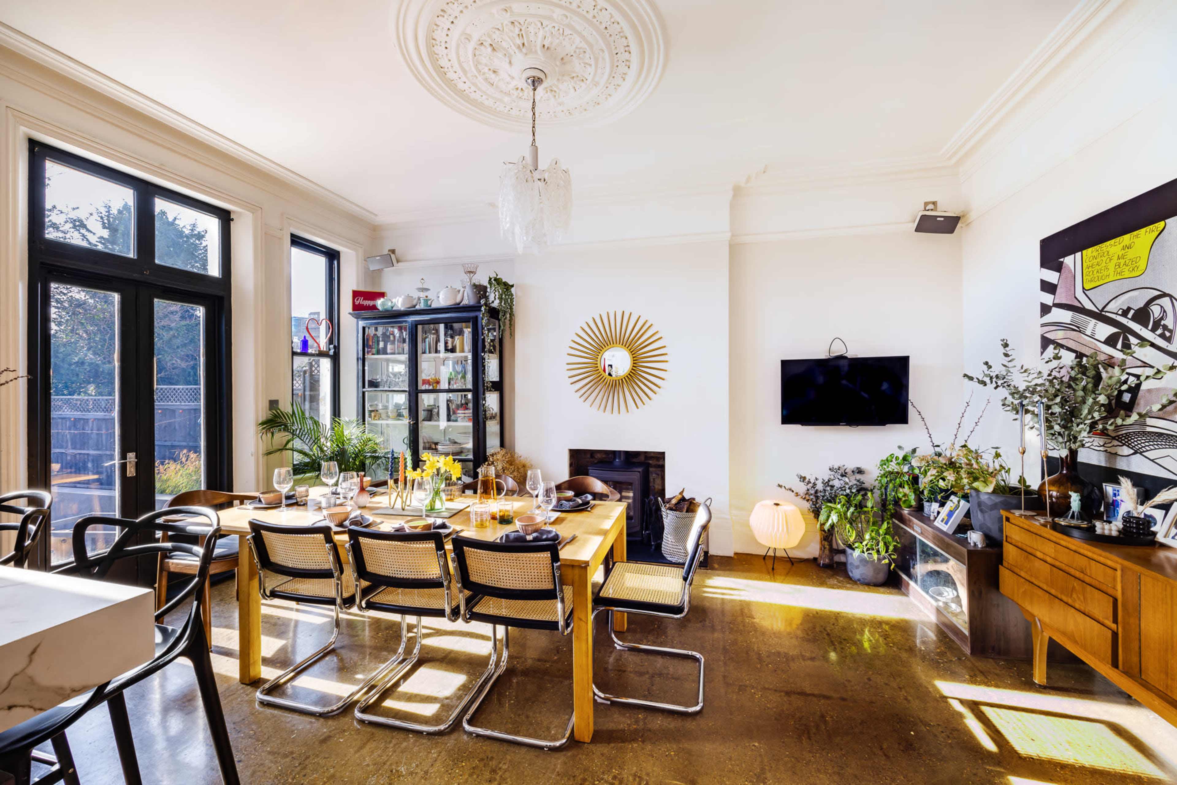 The image shows a bright dining area featuring a large wooden table surrounded by chairs, a wall-mounted television, and various plants alongside a stylish display cabinet.