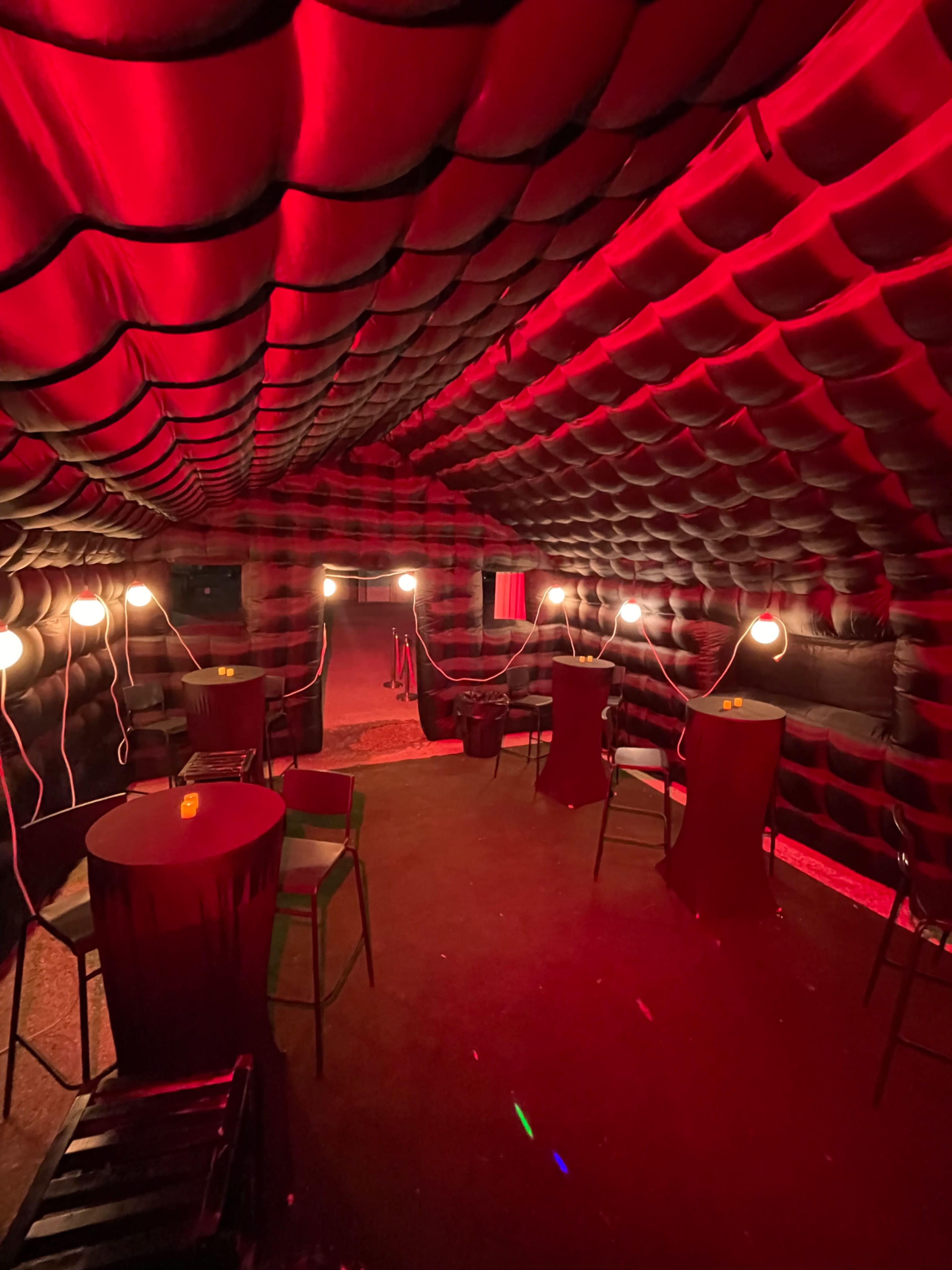 The image shows an interior of a dimly lit, inflatable structure with red lighting, featuring several tables and chairs arranged around the space.