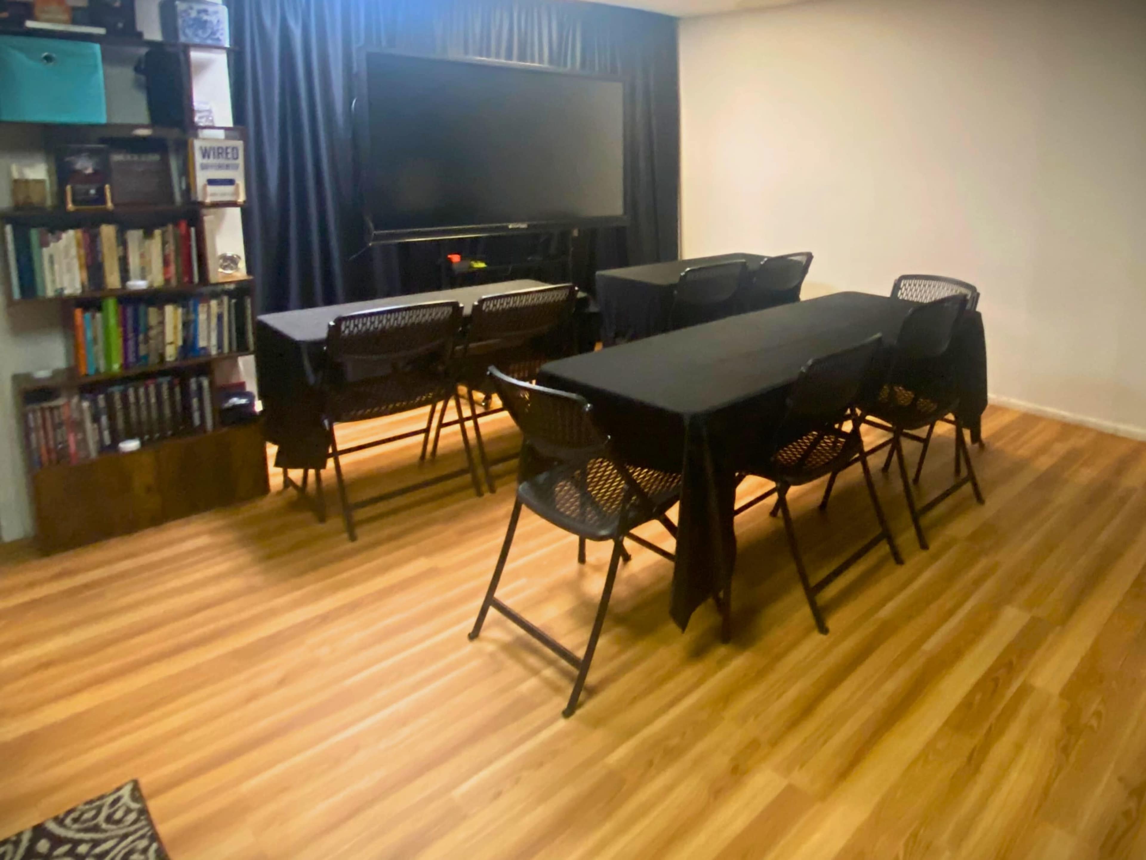 The room features several black folding tables arranged with chairs, a large television on the wall, and a bookshelf in the background.