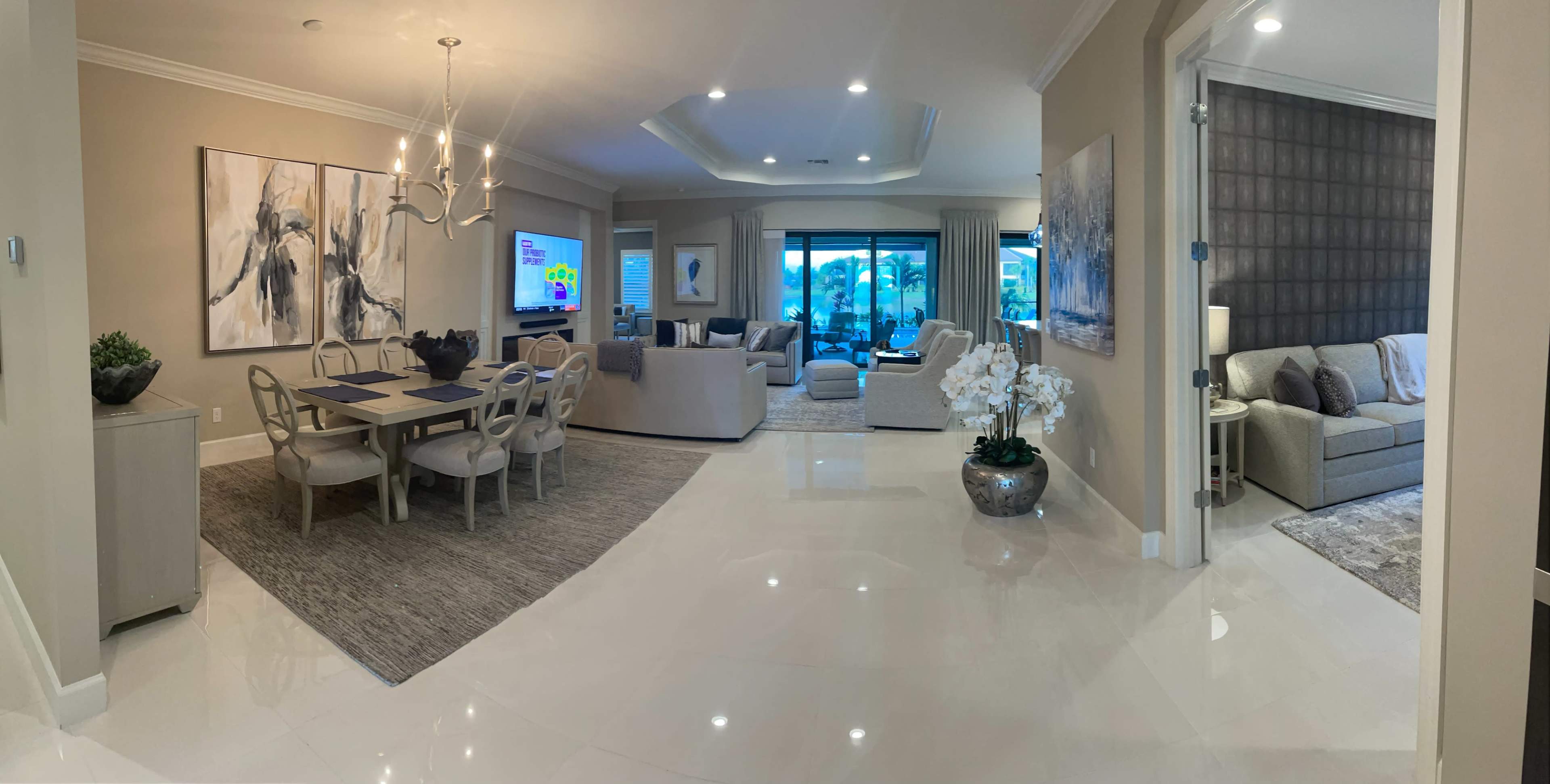 A spacious, elegantly furnished living area with a dining table on the left, a lounge area in the center, and large windows that overlook an outdoor space.