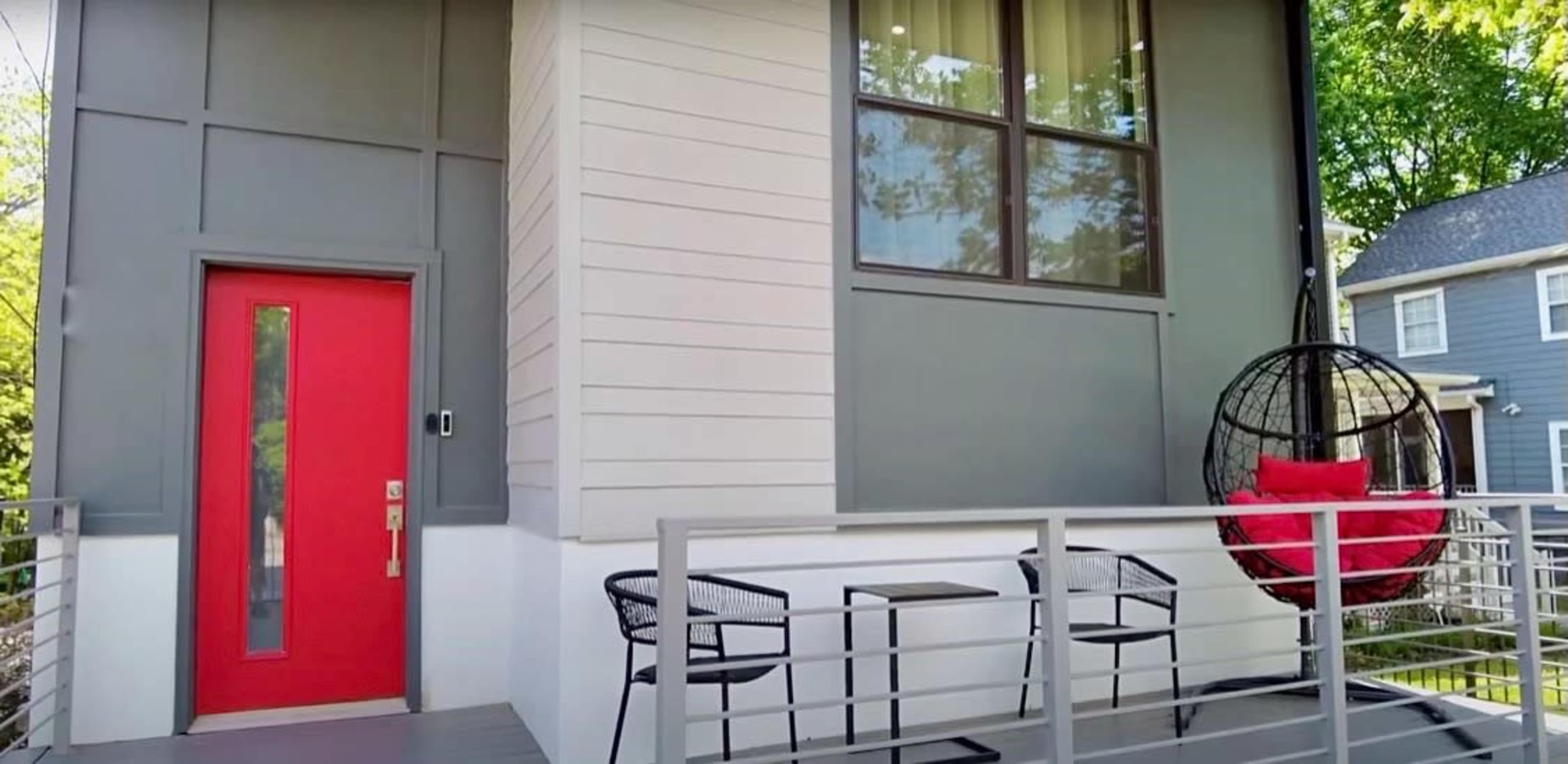 A modern house exterior with a red front door, a hanging chair on the porch, and a railing with two black chairs and small tables.