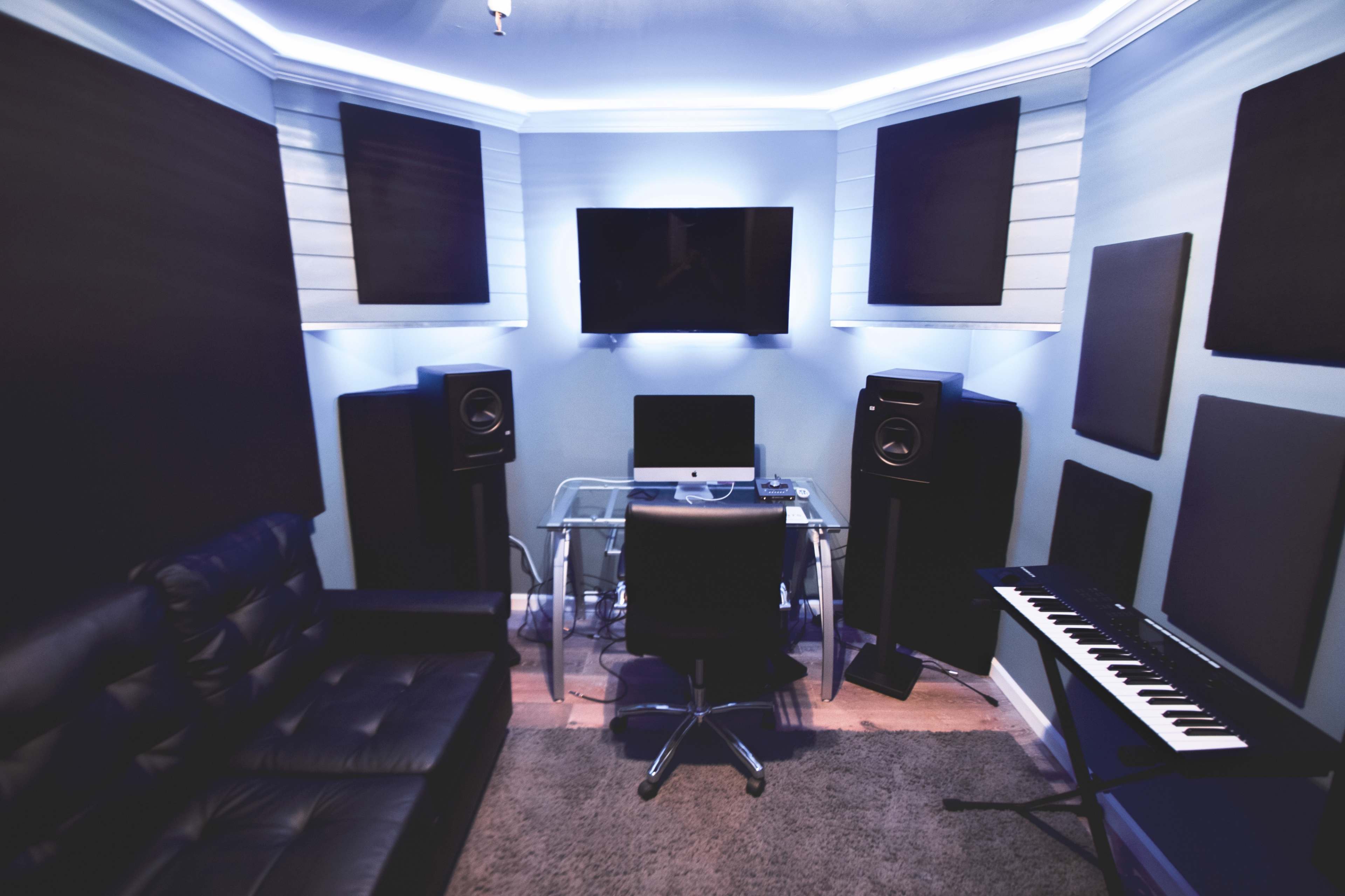 A music production studio featuring a glass desk, an office chair, audio monitors, and a keyboard, with soundproofing panels on the walls and a television mounted above.