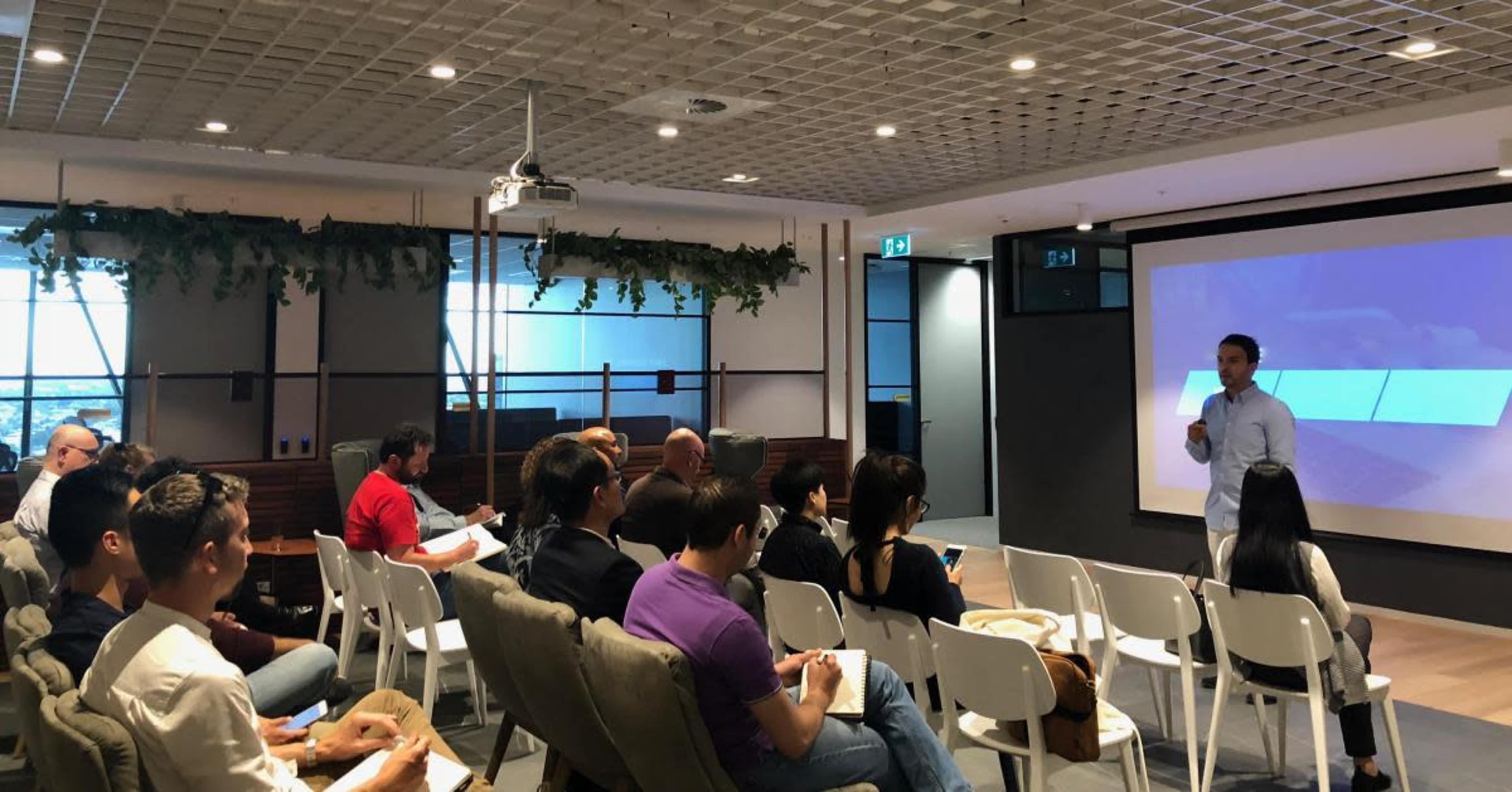 A speaker presents in front of an audience seated in a modern conference room with large windows and a projector screen.
