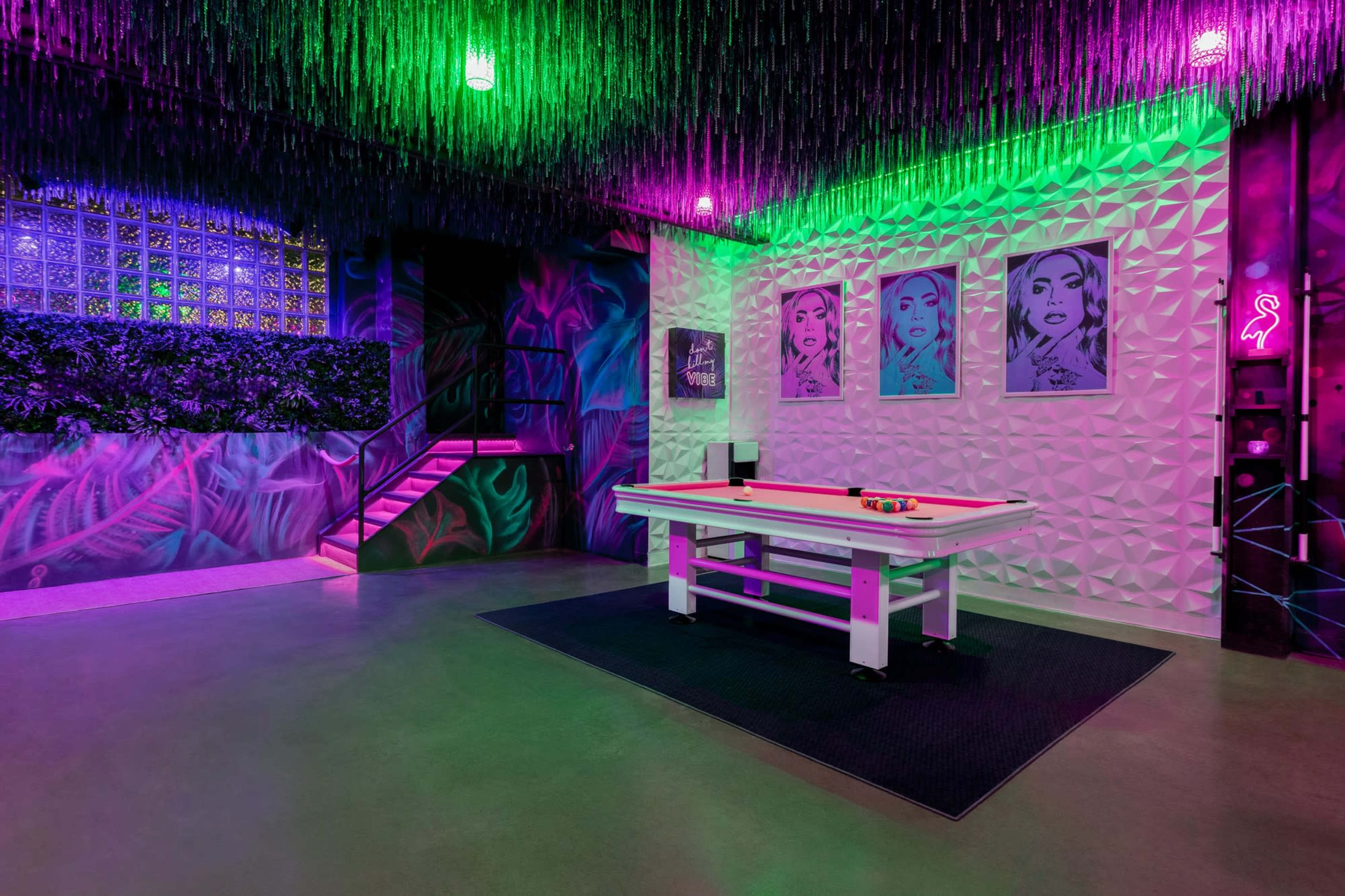 A vibrant game room featuring a pool table, colorful neon lighting, and bold wall art with framed portraits.