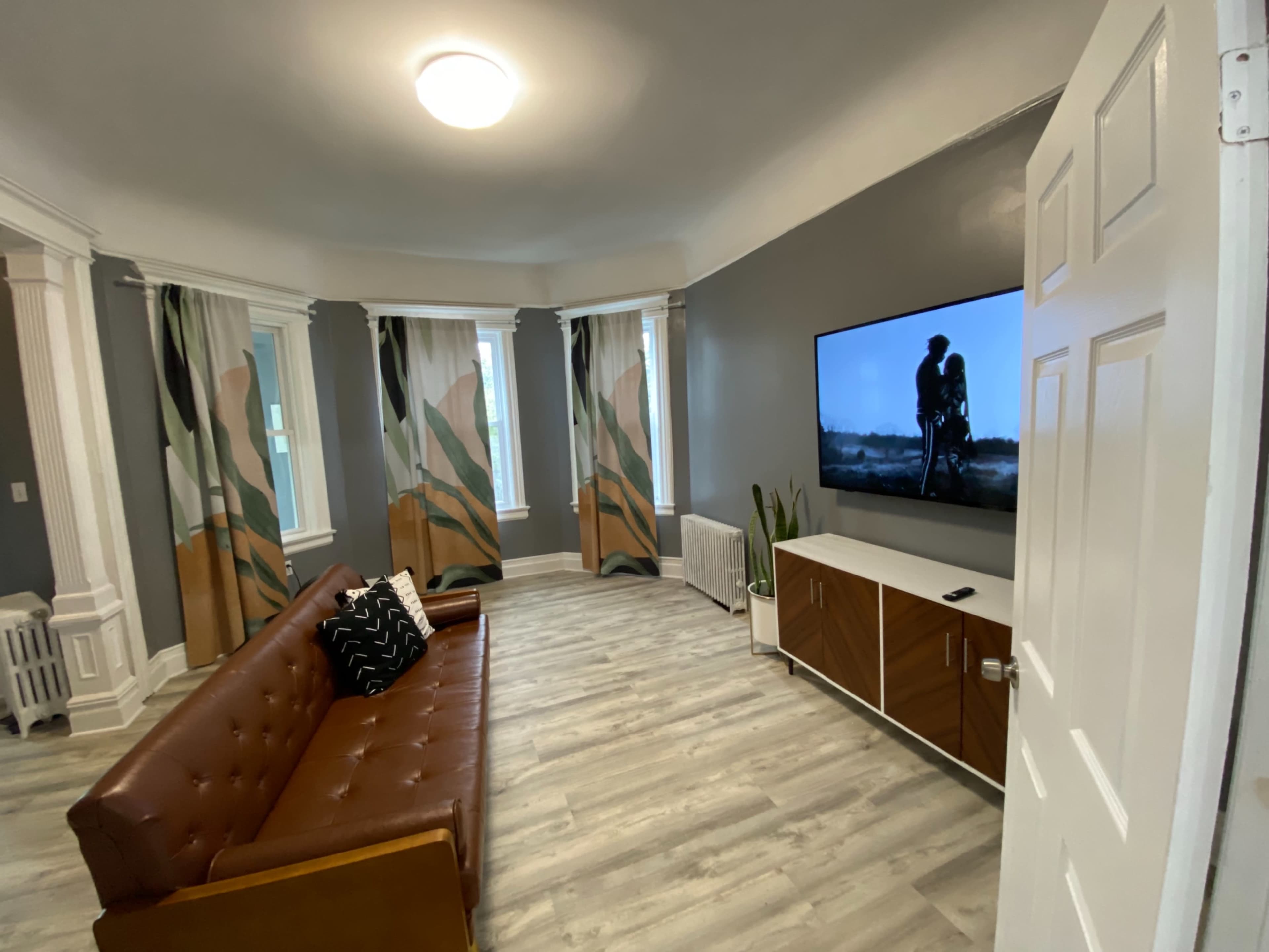 A modern living room features a brown leather couch, a wall-mounted television displaying an image, and large windows draped with patterned curtains.