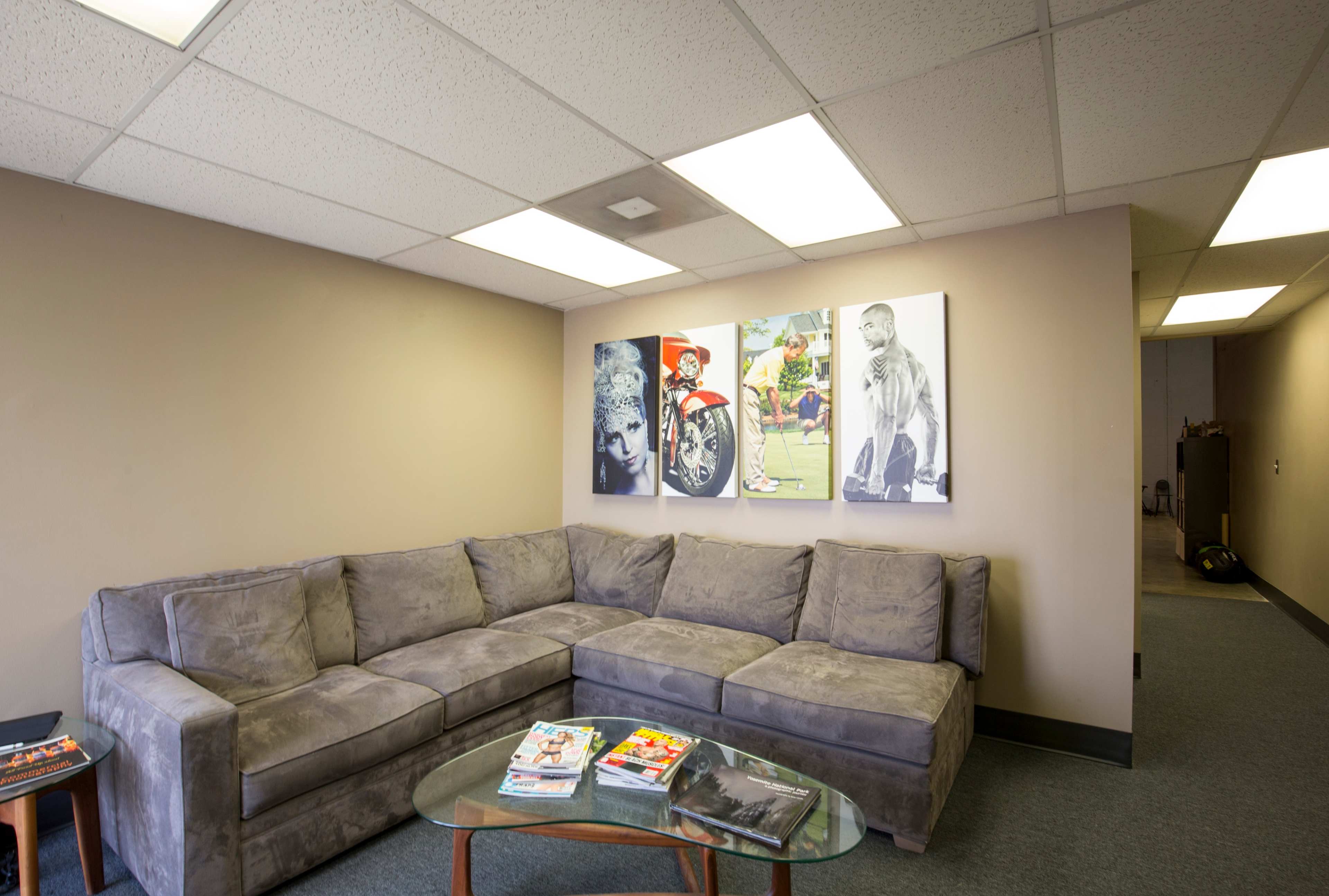 The image shows a cozy lounge area featuring a gray sectional sofa, a glass coffee table with magazines, and three large framed pictures on the wall.