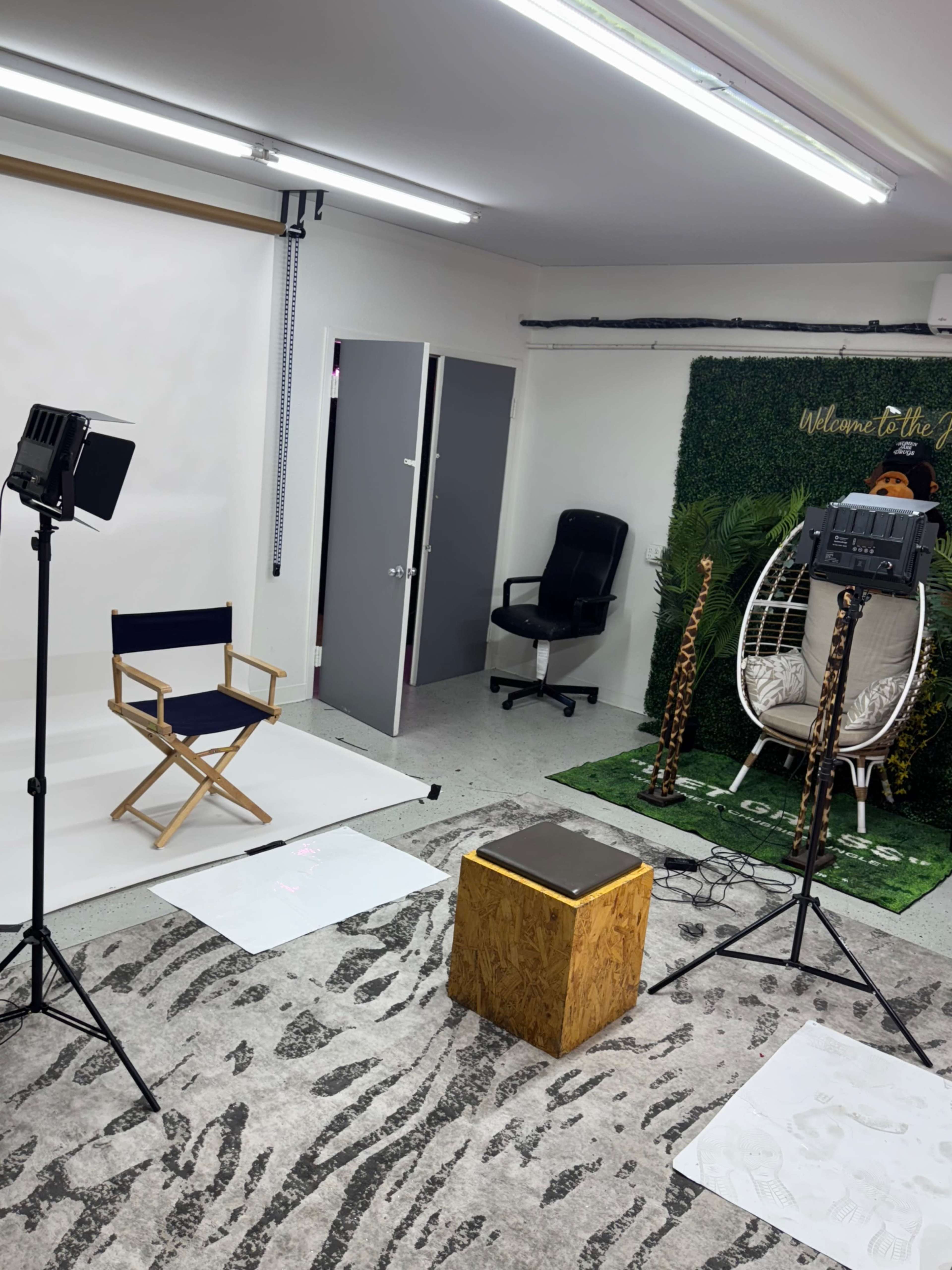 The image shows a photography or video studio setup with a director's chair, lighting equipment, a small wooden stool, and a background featuring greenery and a decorative sign.