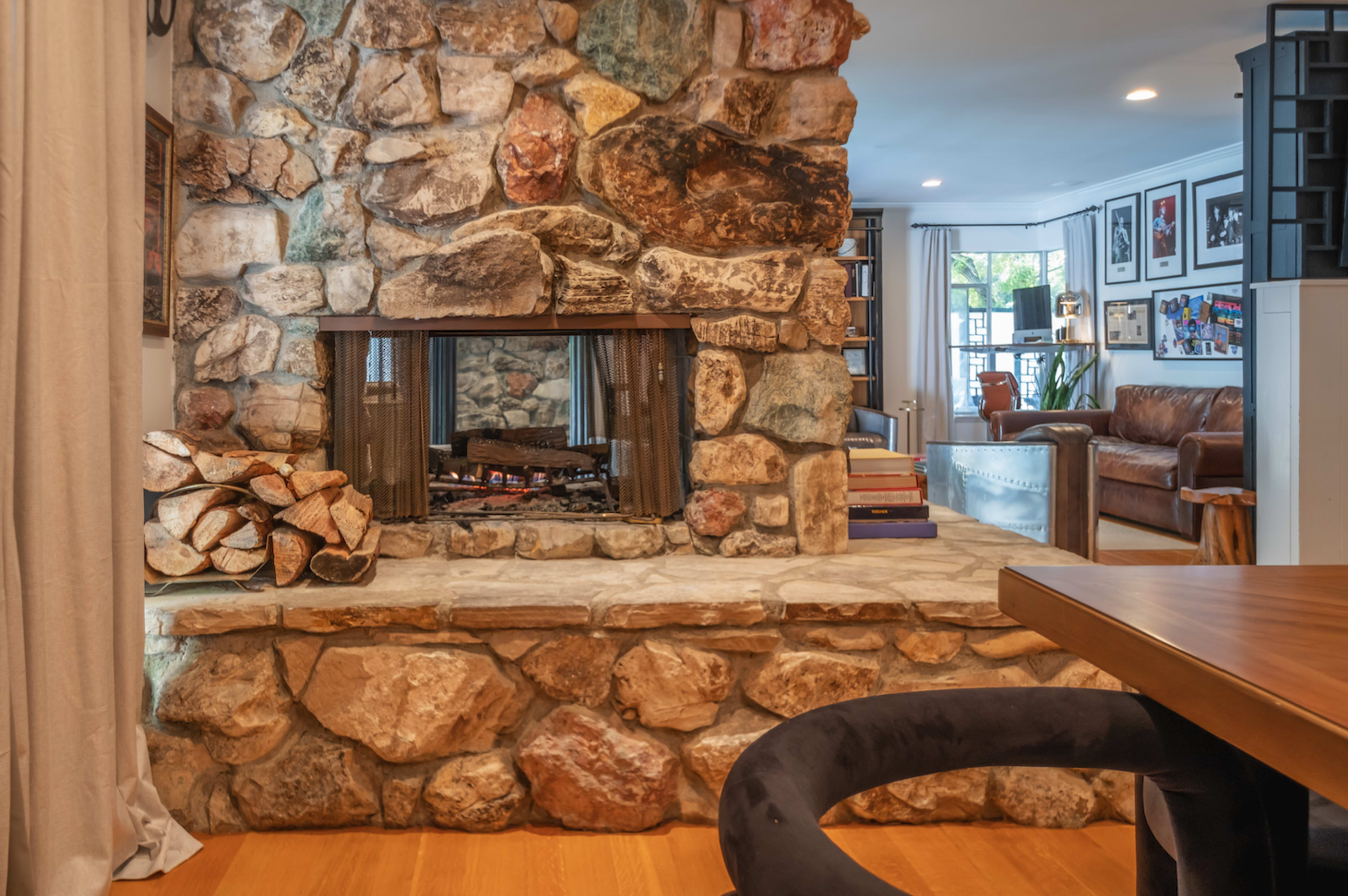 A stone fireplace set in a living room with a wooden floor, a leather sofa, and a stack of logs beside it.