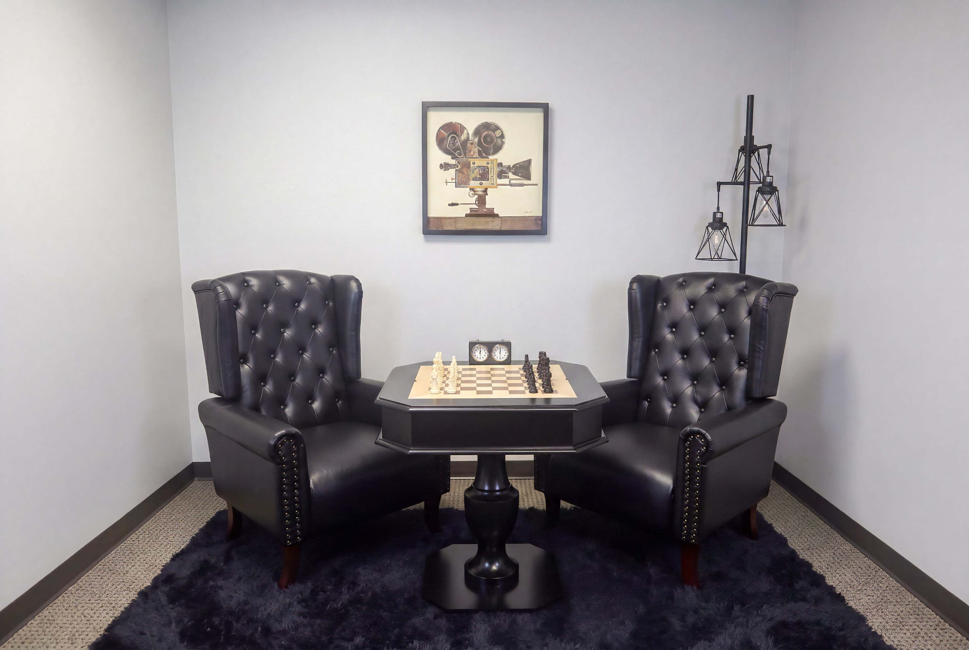 The image shows a small gaming area with two black leather armchairs facing a chessboard on a table, accompanied by a decorative artwork on the wall.