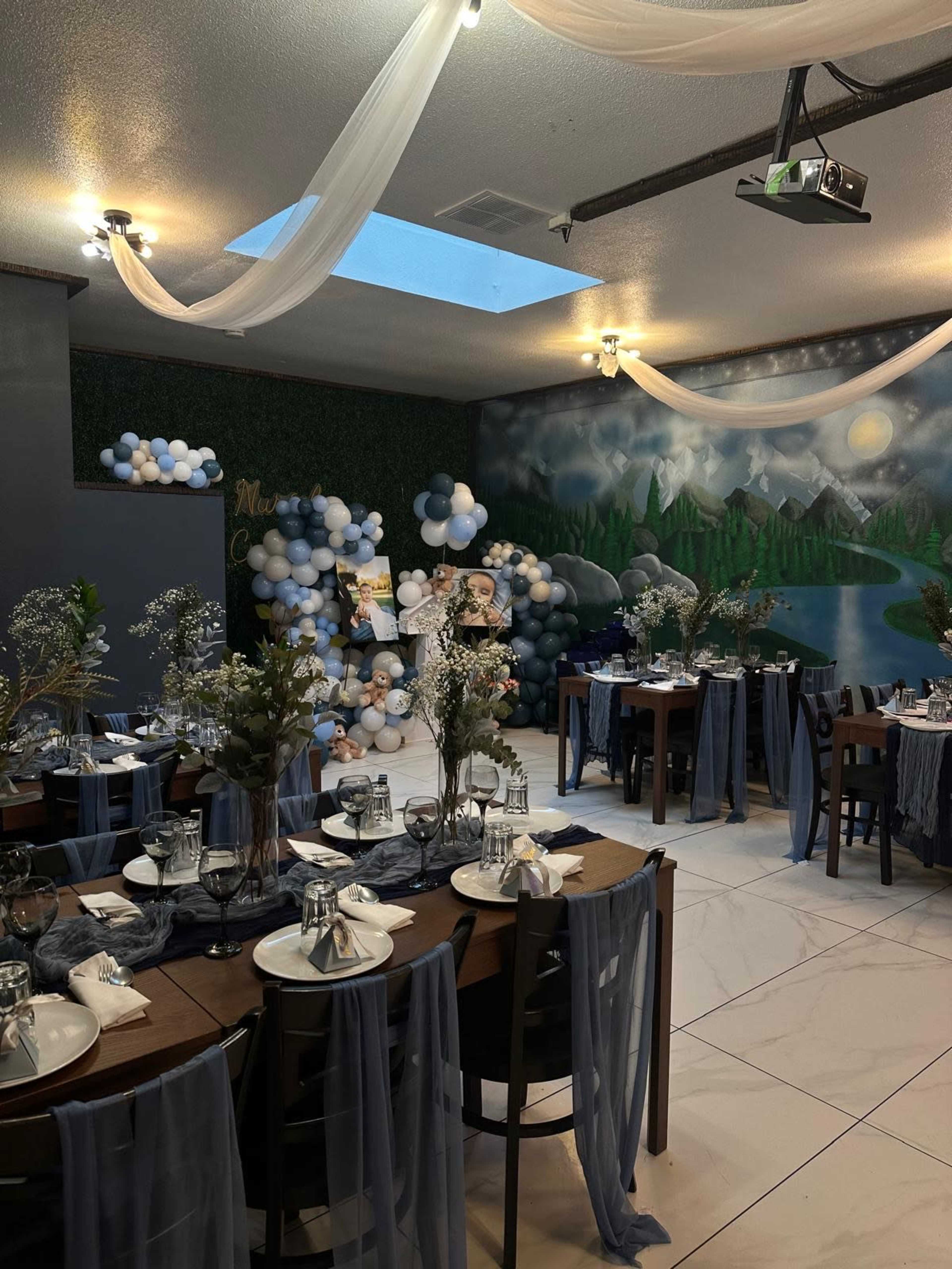 The image shows a decorated event space with tables set for dining, blue and white balloon arrangements, and a mural of a lake surrounded by mountains.
