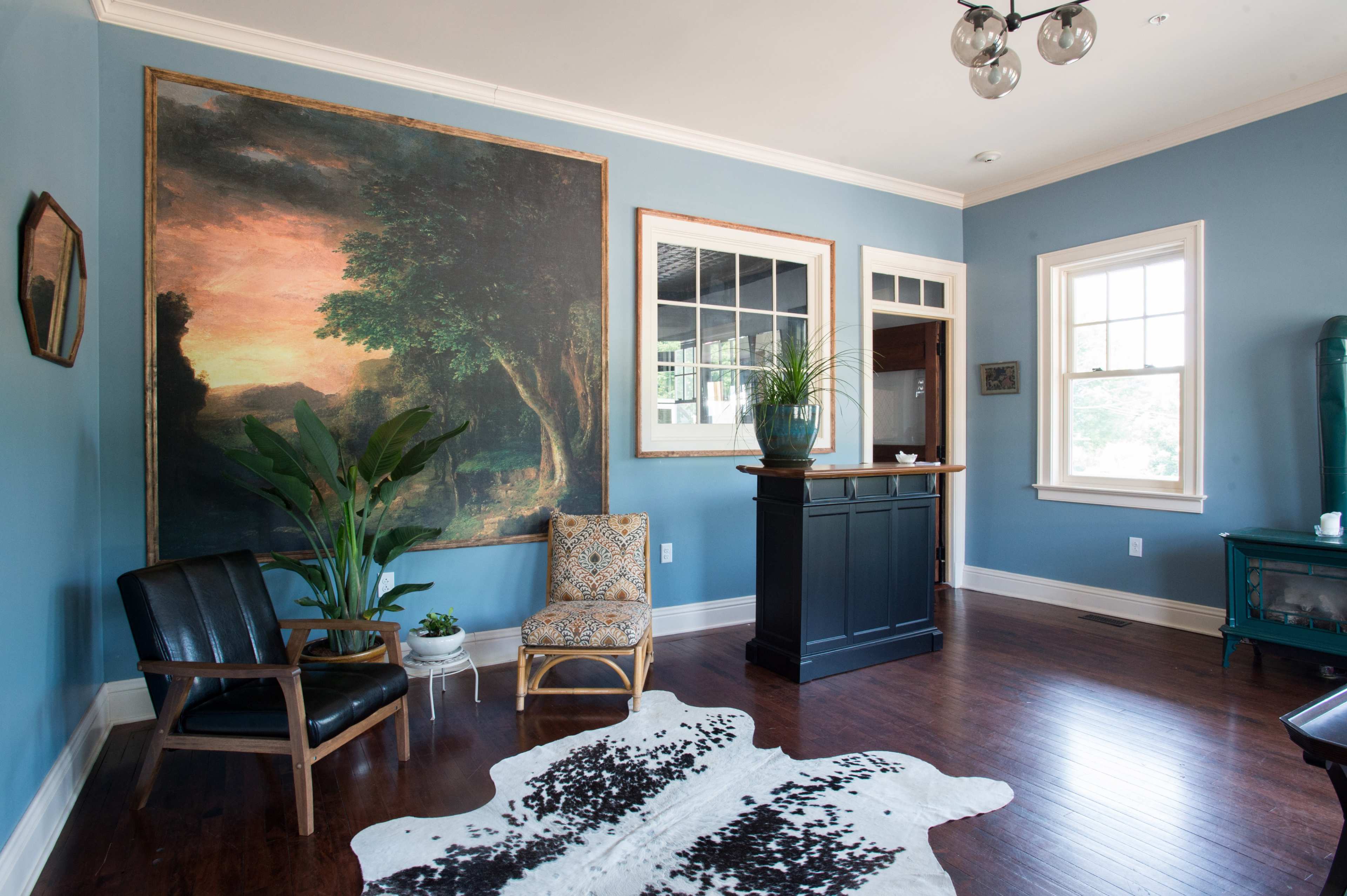 The room features blue walls, a large landscape painting, a patterned armchair, a sleek black chair, and a small bar area with plants and natural light.