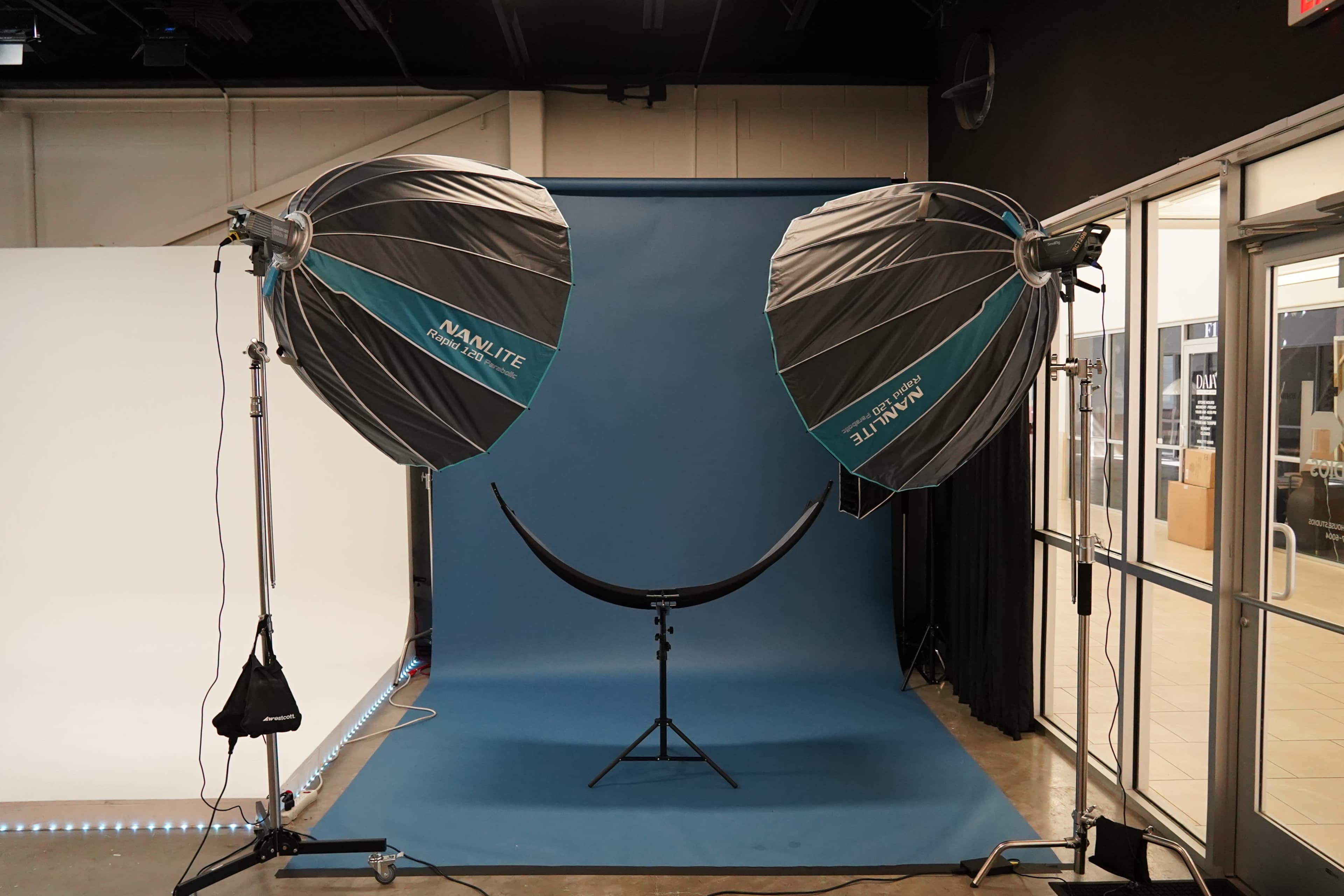 The image shows a photography studio setup with two large softbox light fixtures positioned at the front, directed toward a curved backdrop in a light blue color.
