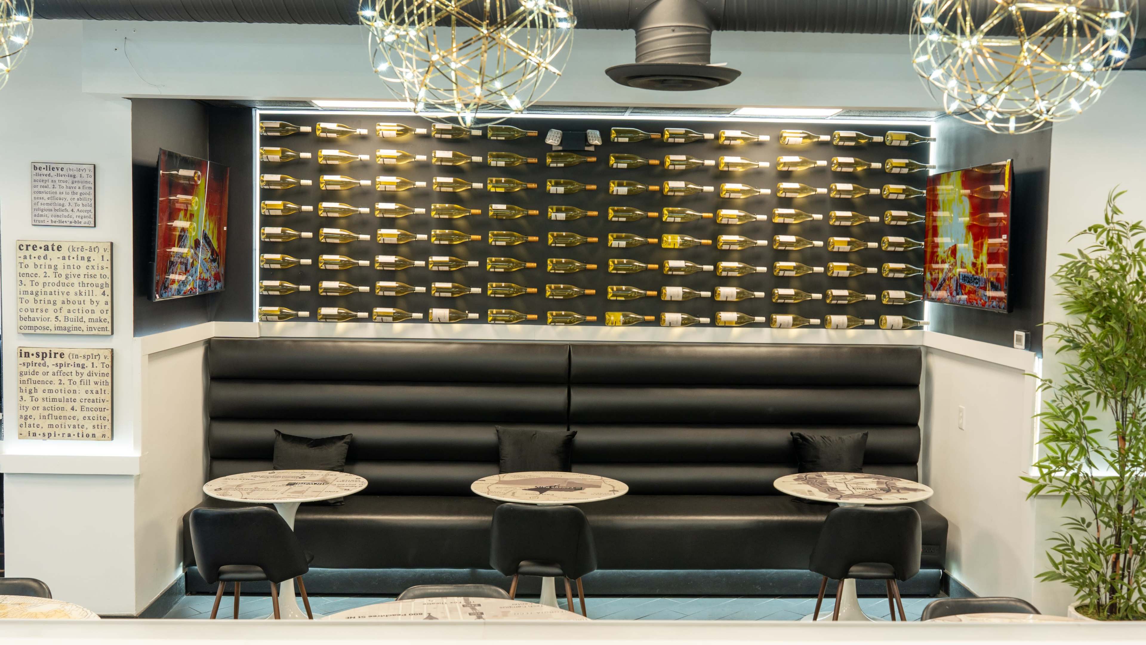 The image shows a modern seating area in a bar or restaurant with a black cushioned booth, circular tables, and decorative wall-mounted displays of champagne bottles.