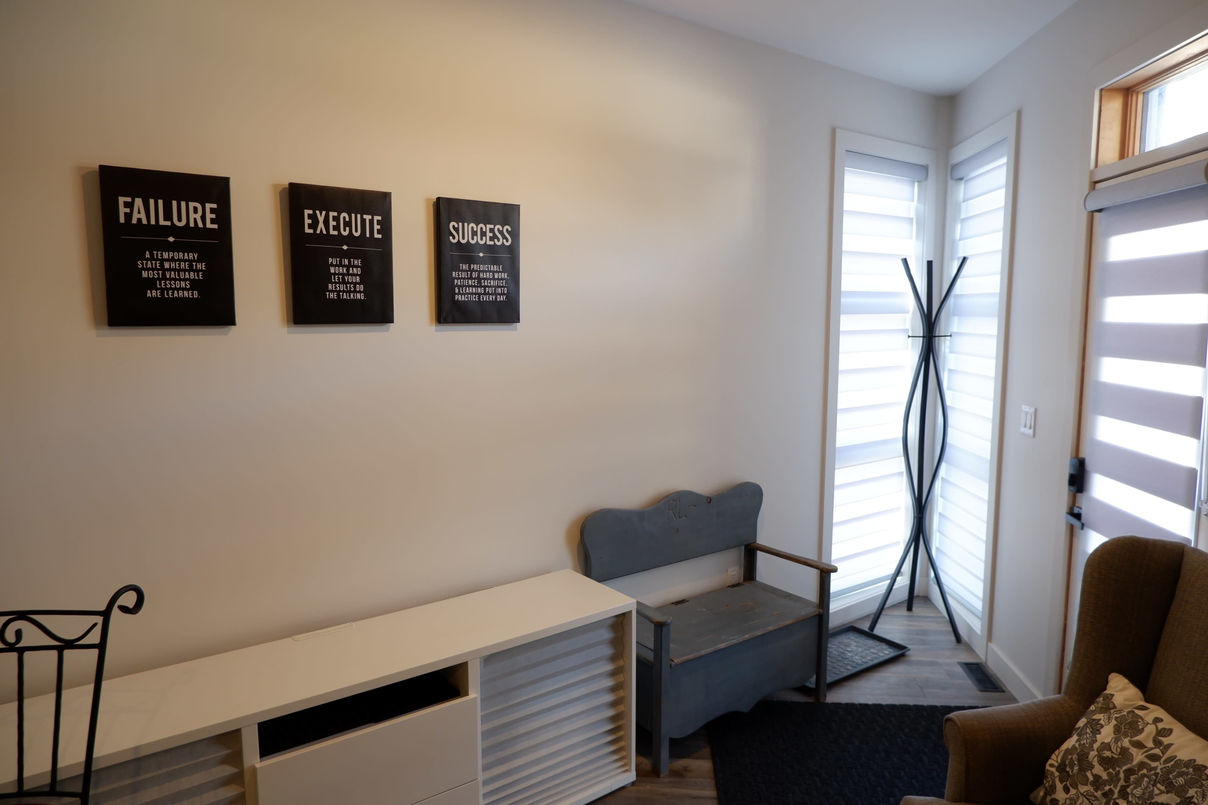The image shows a minimalist interior space featuring three framed motivational quotes on the wall, a wooden bench, and a coat rack near a window with individually adjustable slats.