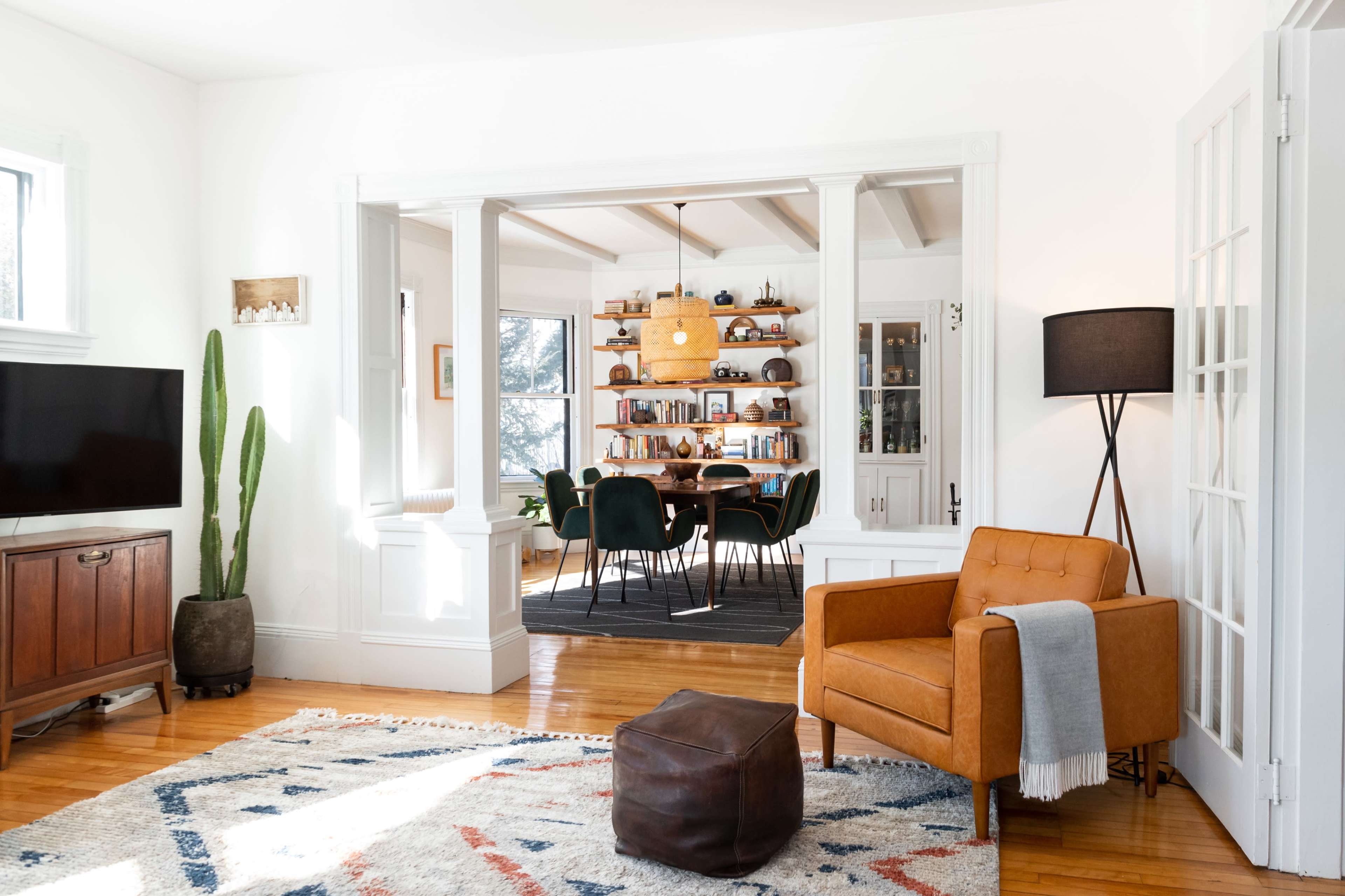 A bright living room featuring an orange armchair, a decorative rug, a television, and a view into a dining area with a wooden table and bookshelves.