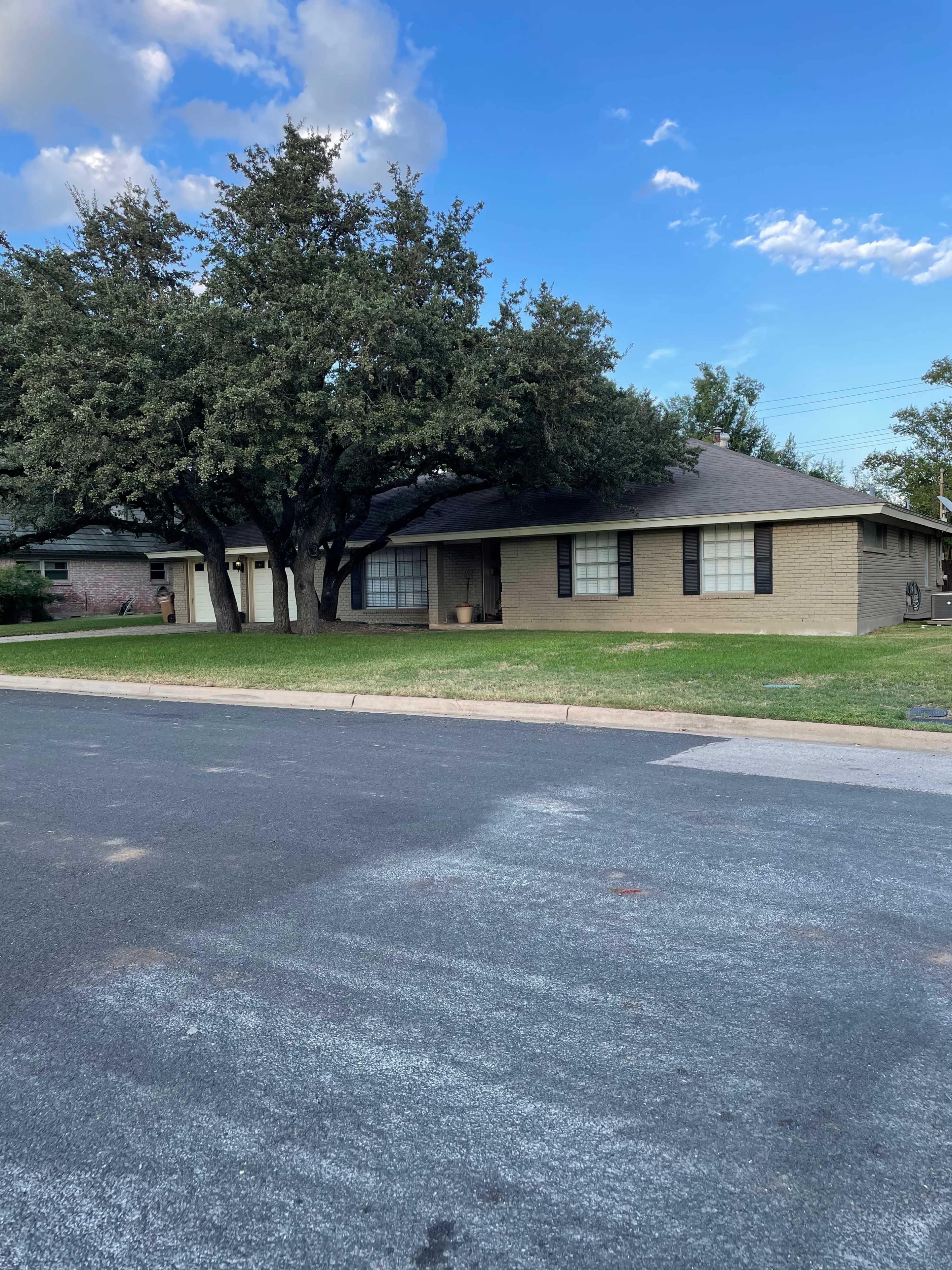 A single-story house with a brick exterior and large trees in the front yard is situated along a quiet street.