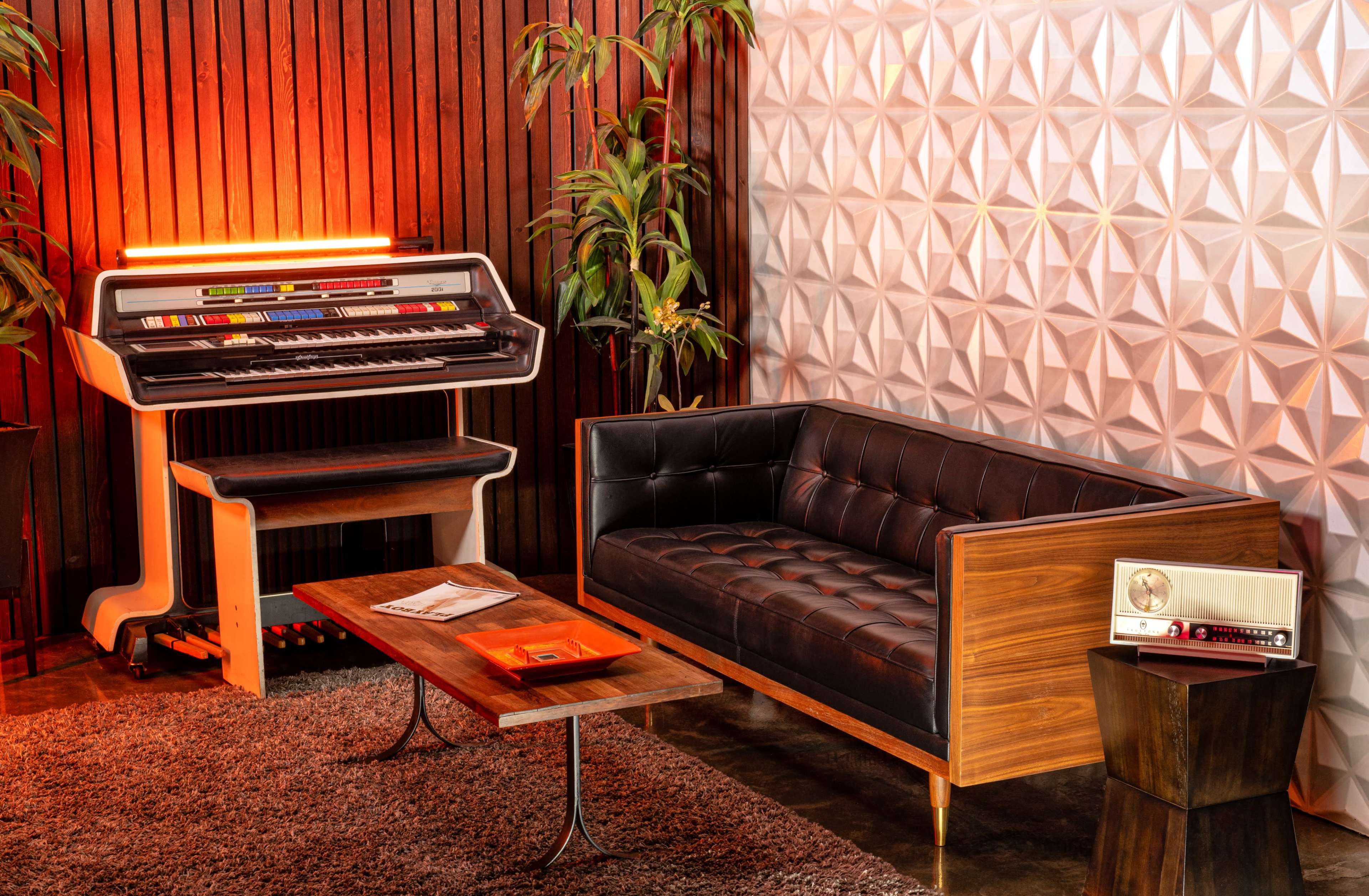 The image features a modern living space with a black leather couch, a wooden coffee table, a vintage radio, and a colorful keyboard instrument against a backdrop of textured wall panels and indoor plants.