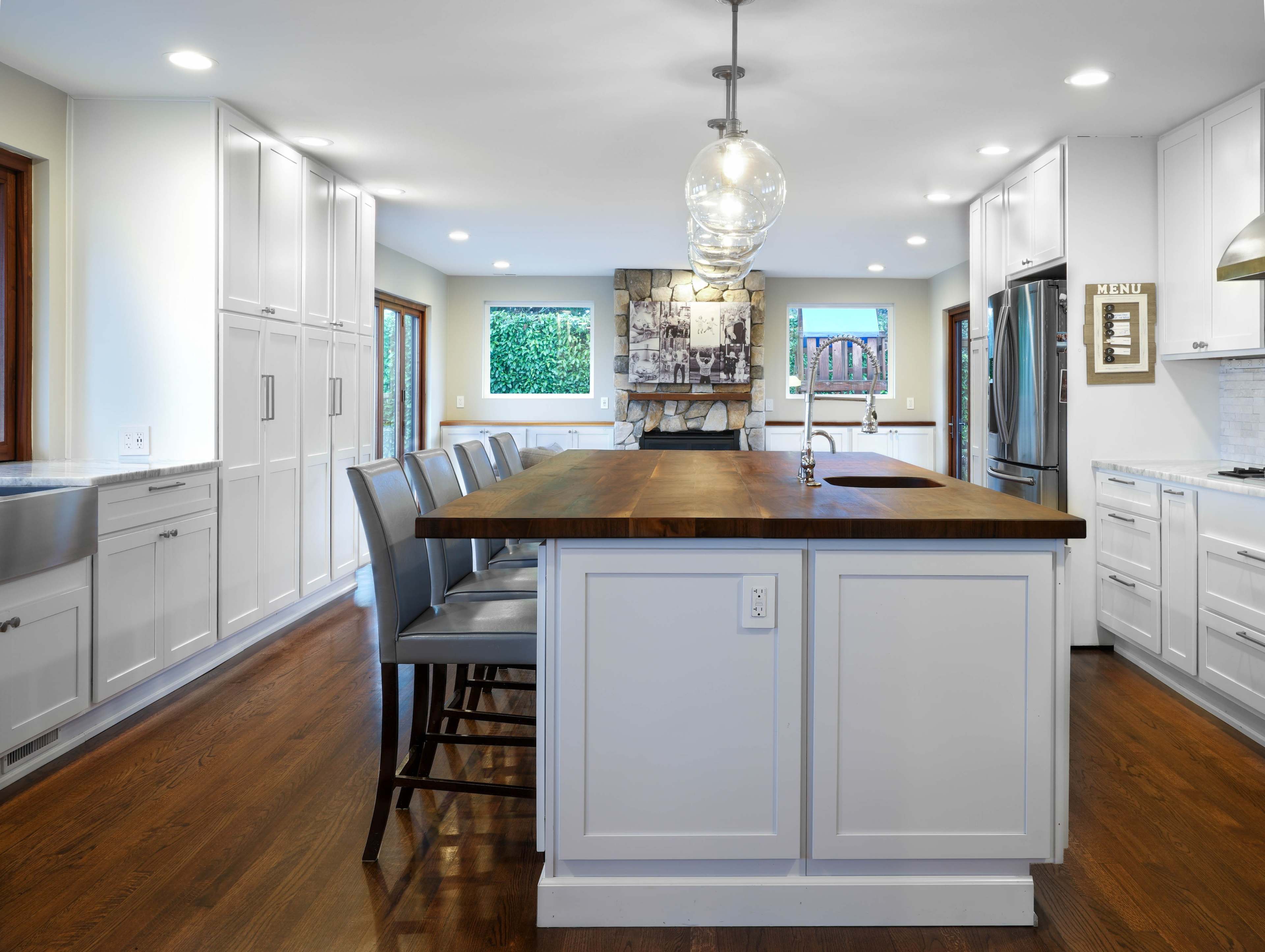 A modern kitchen features a central wooden island with seating, white cabinetry, and large windows that provide natural light.