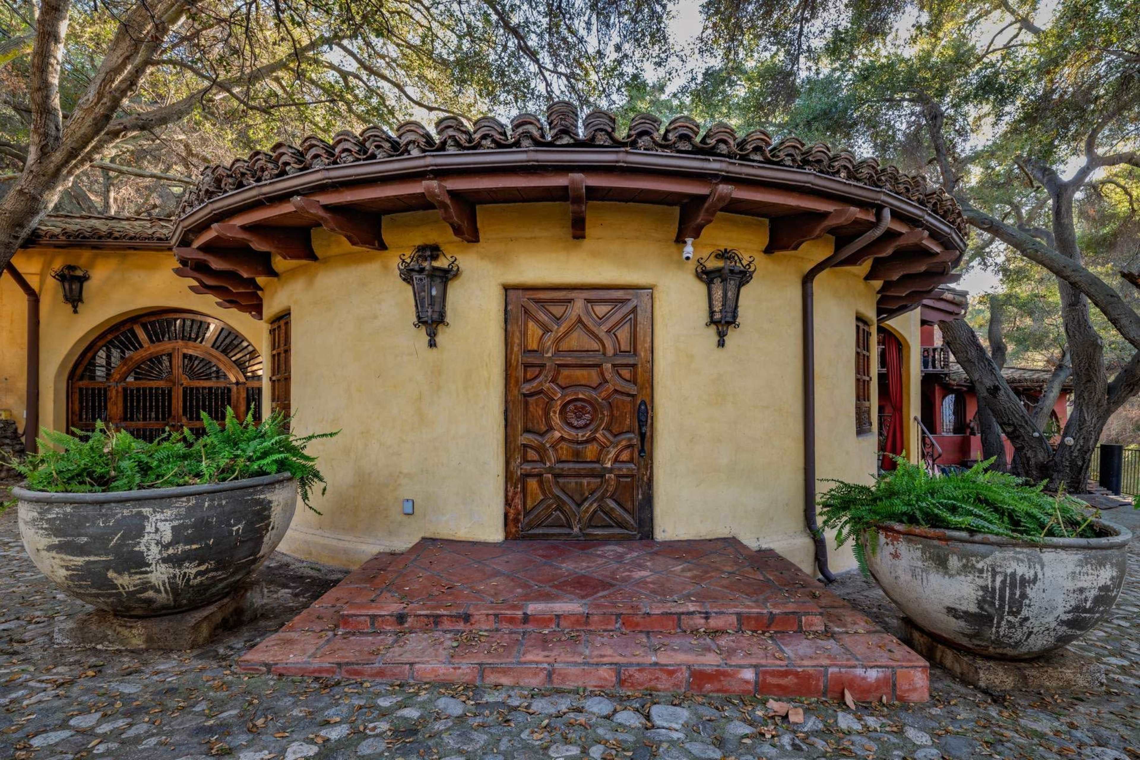 The image depicts a round, yellow stucco building with a wooden door and decorative light fixtures, surrounded by large planters and nestled among trees.