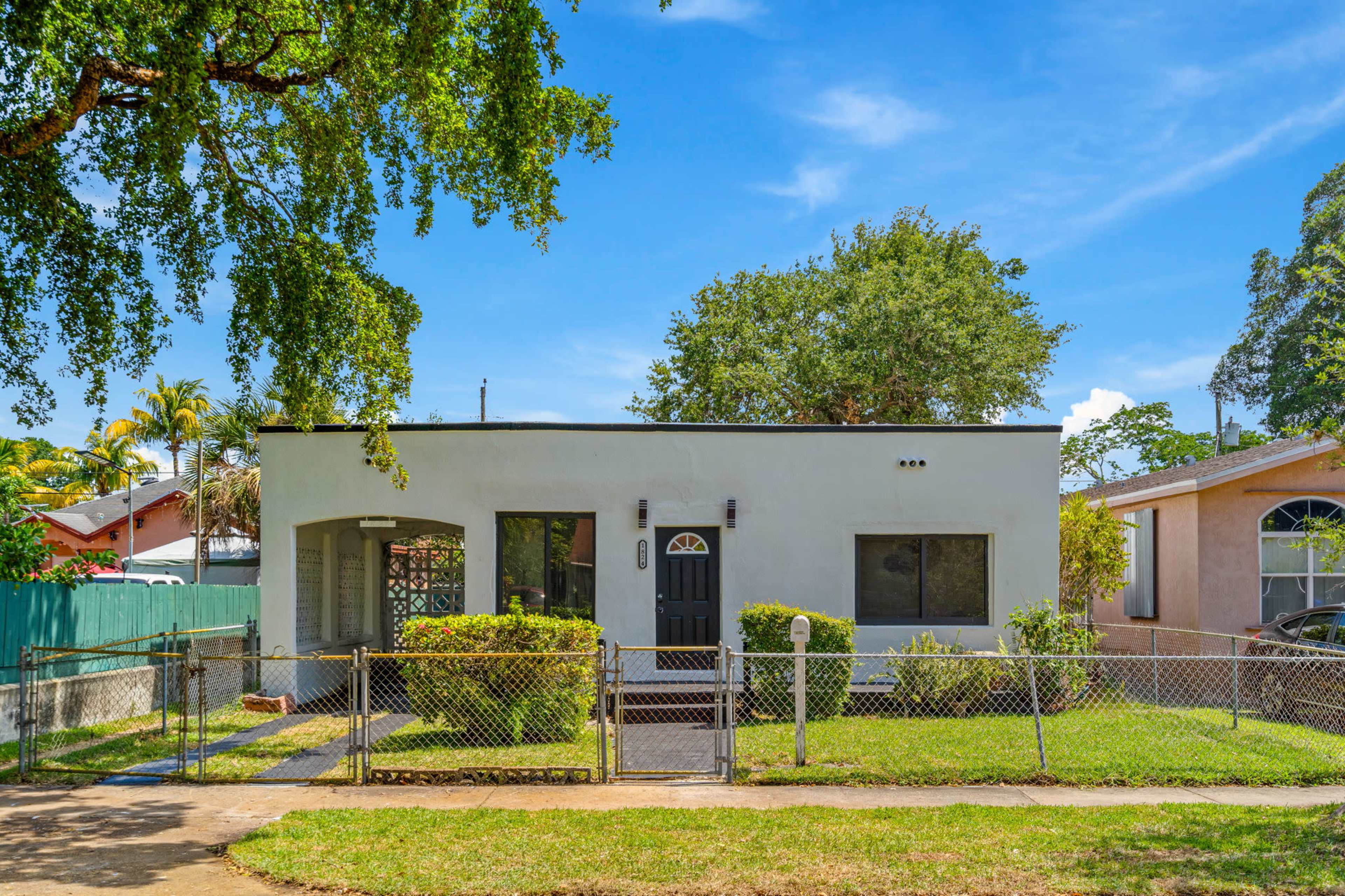 A single-story house with a black front door and a fenced yard is situated under a large tree, surrounded by green grass and neighboring homes.