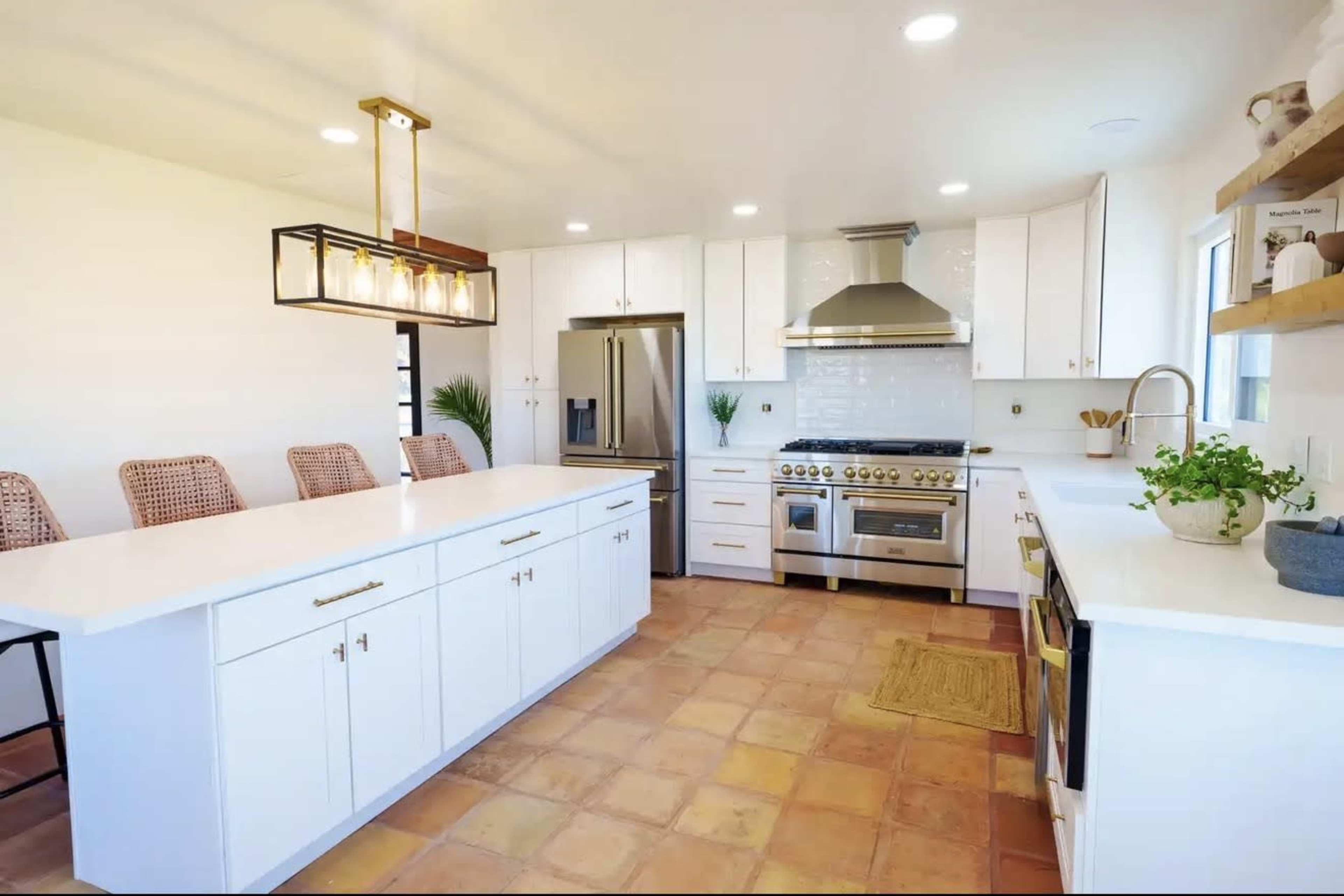 The image shows a modern kitchen with white cabinetry, a large island with seating, stainless steel appliances, and terracotta tile flooring.