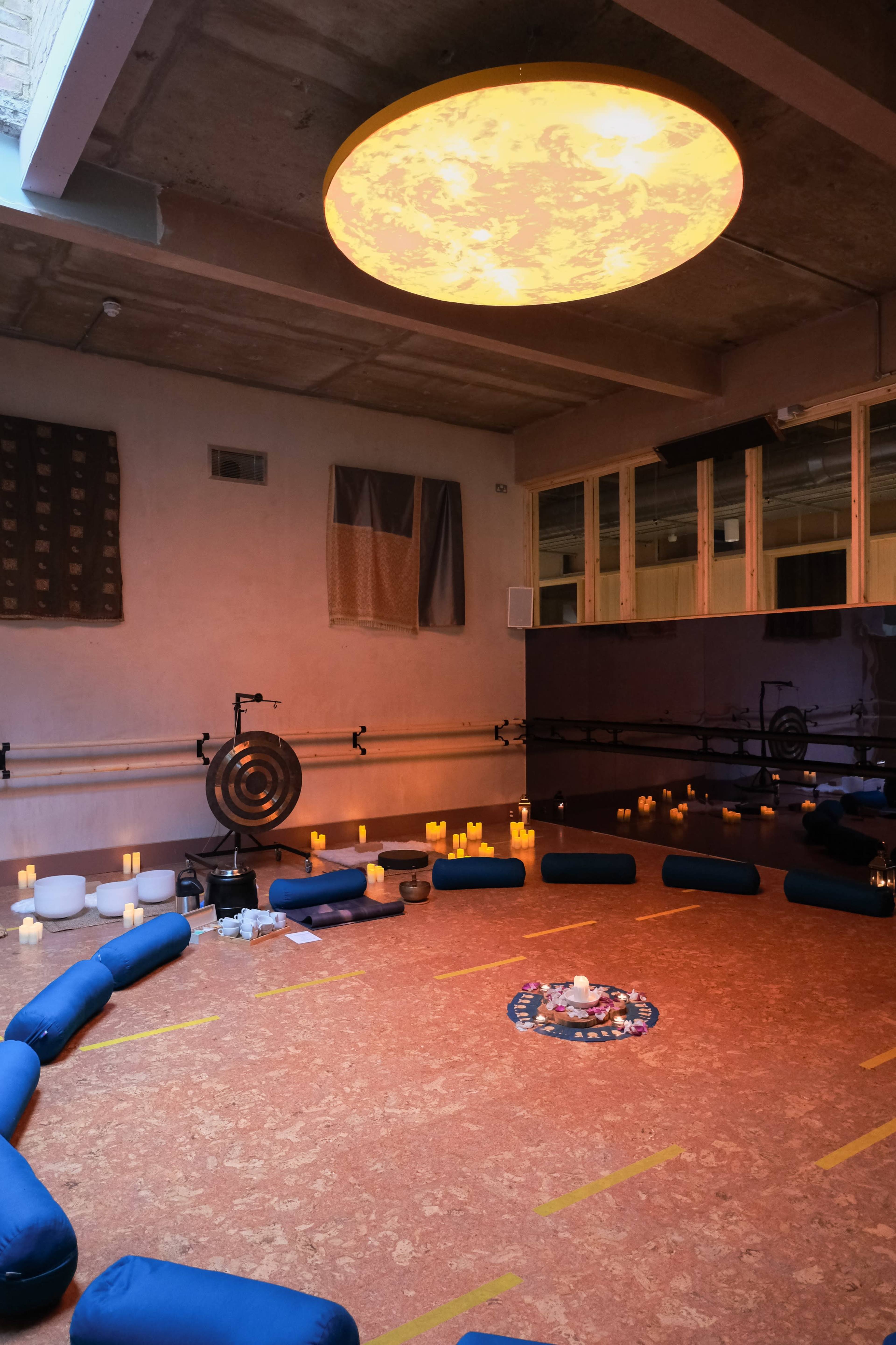 The image shows a serene meditation room with a circular arrangement of cushions, candles, and a large illuminated ceiling fixture.
