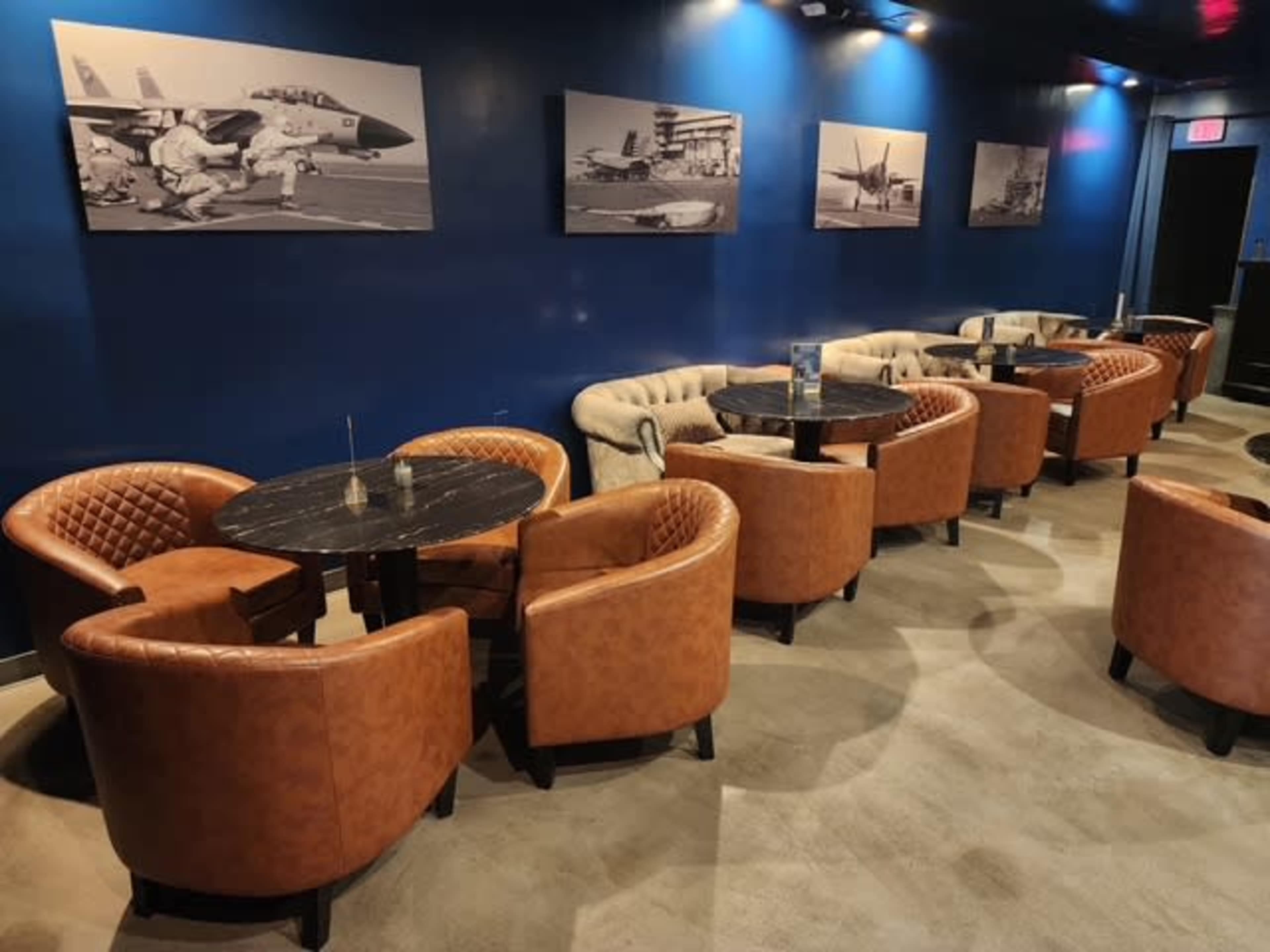 The image shows a stylish lounge area with leather armchairs and round marble tables, featuring black and white photographs of aircraft on the walls.
