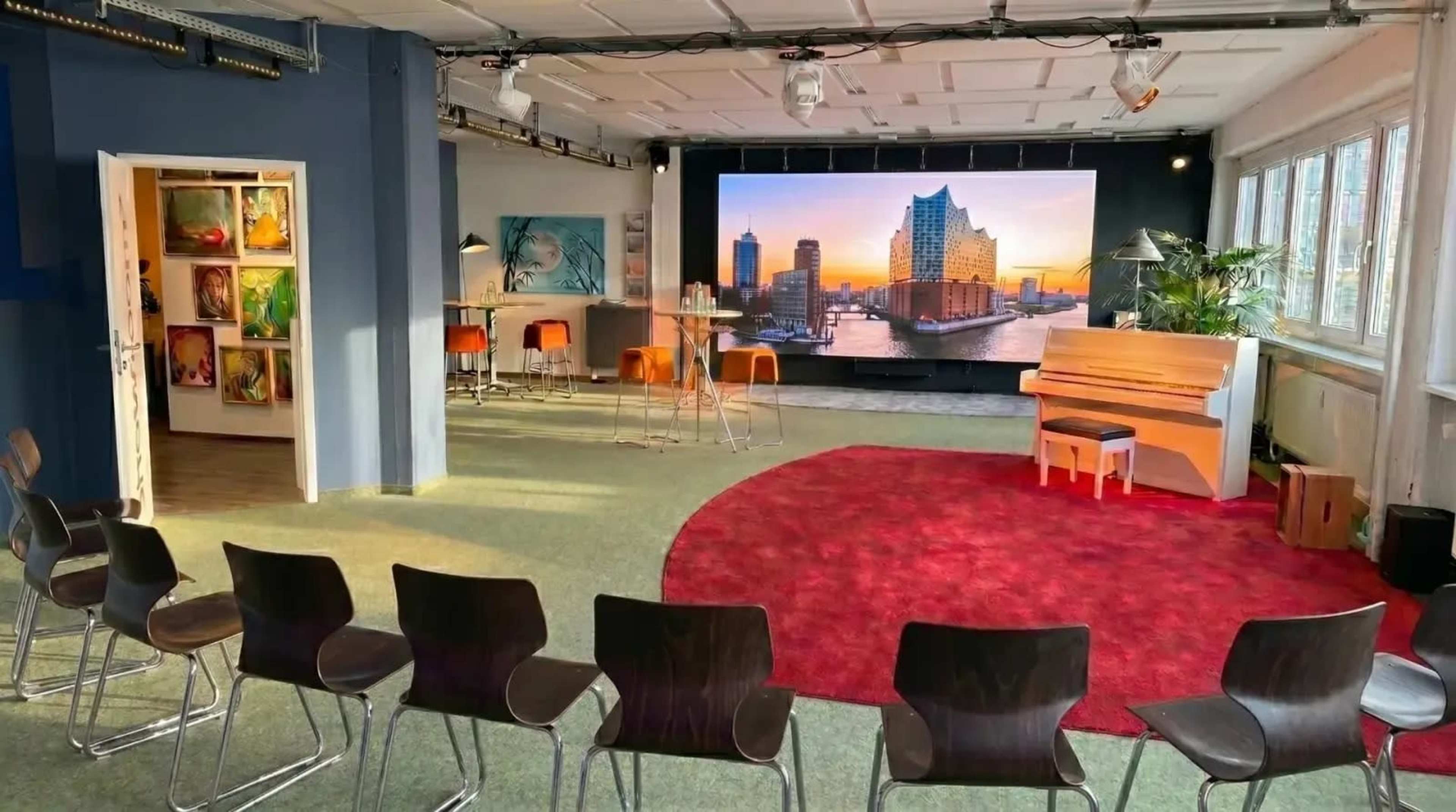 The image shows an indoor event space featuring a red carpet area, a piano, and seating arranged in a semi-circle facing a large screen displaying a cityscape.