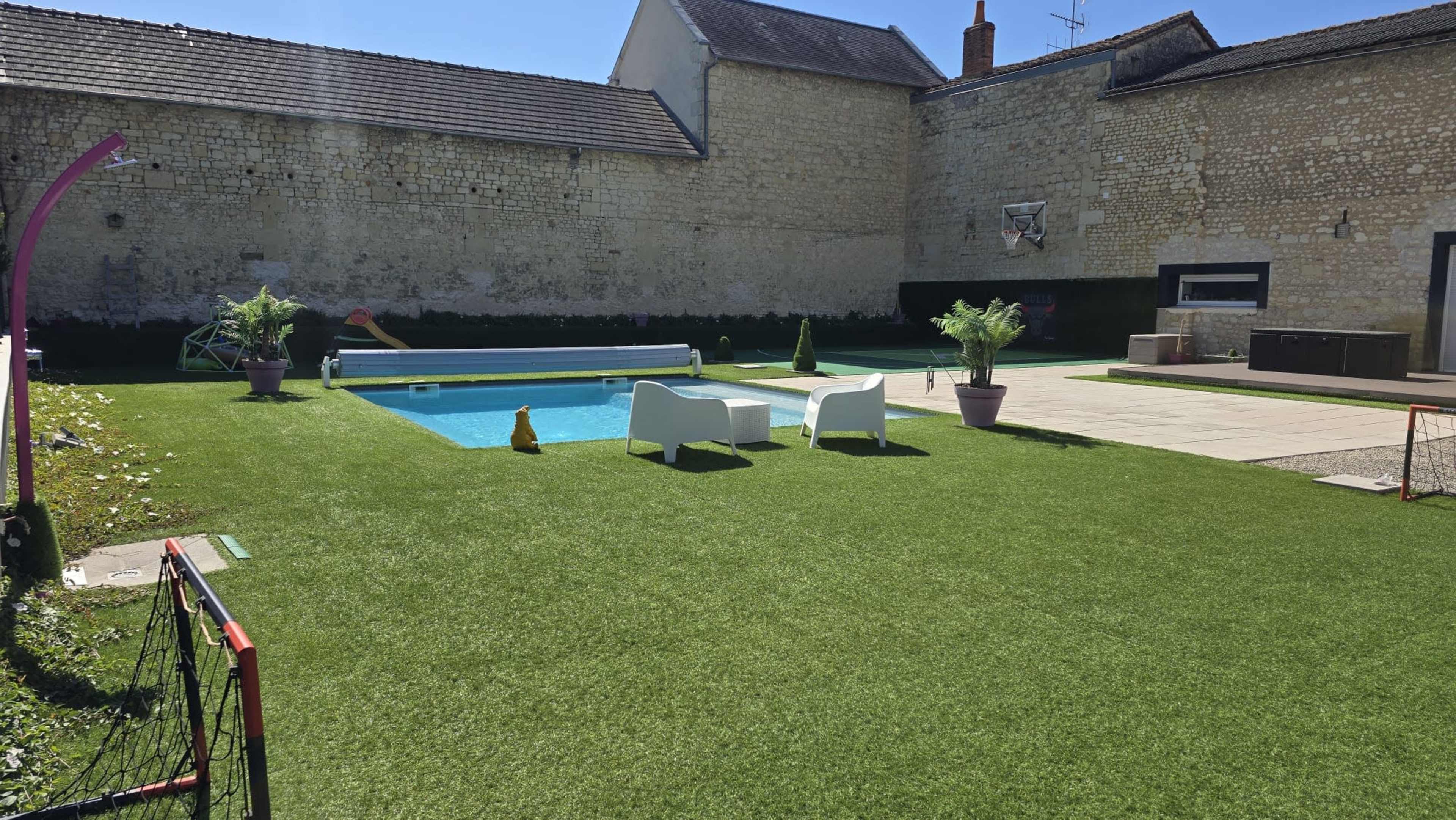 The image shows a backyard with a swimming pool, two white chairs, manicured grass, and a stone wall in the background.