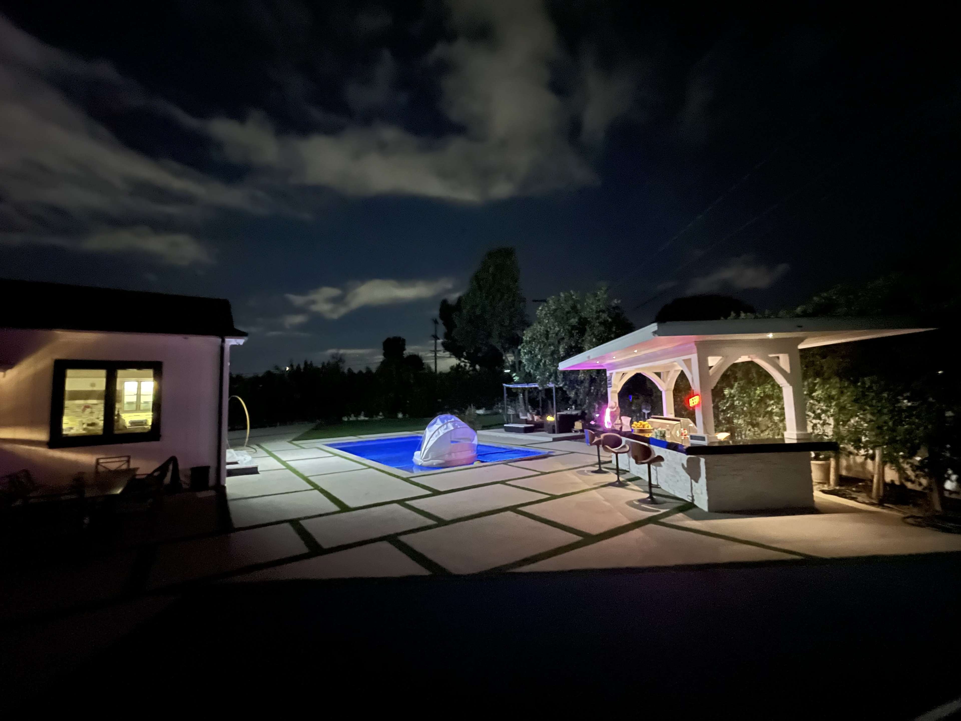 A lit pool area with a bar and seating is visible at night, surrounded by trees and a house with glowing windows.