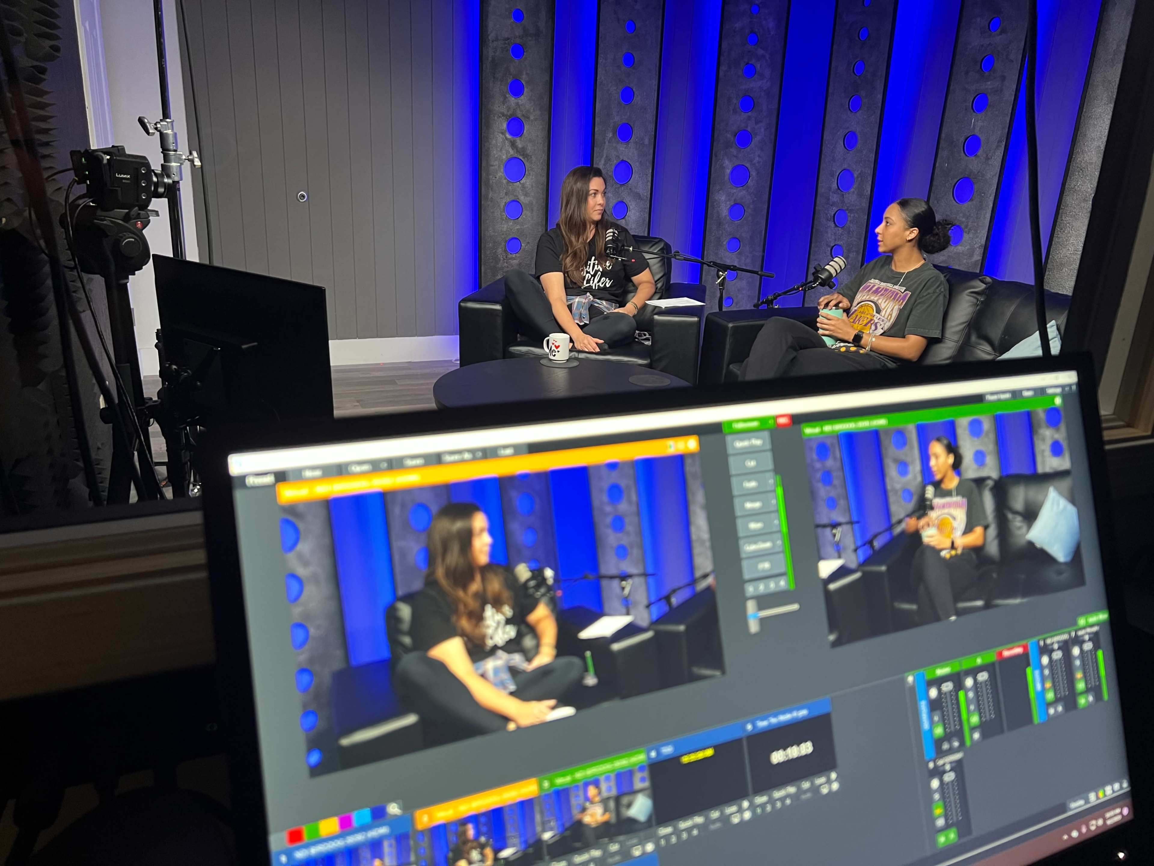 Two people are sitting on a couch in a recording studio, engaged in conversation, with cameras and equipment visible in the background.