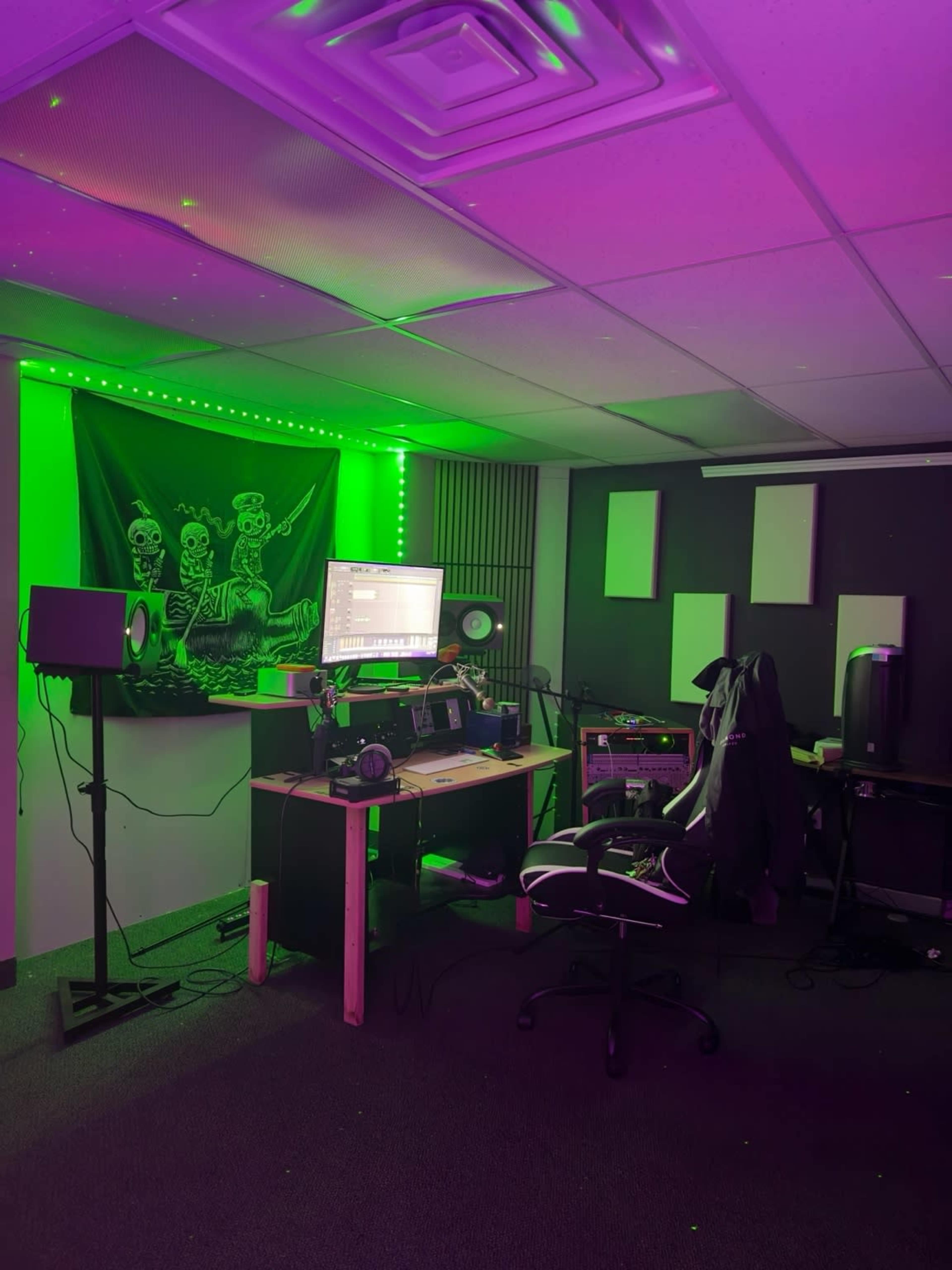 A music production studio features a desk with audio equipment, speakers, and a green-lit wall adorned with a flag, set against a backdrop of dark-colored walls.