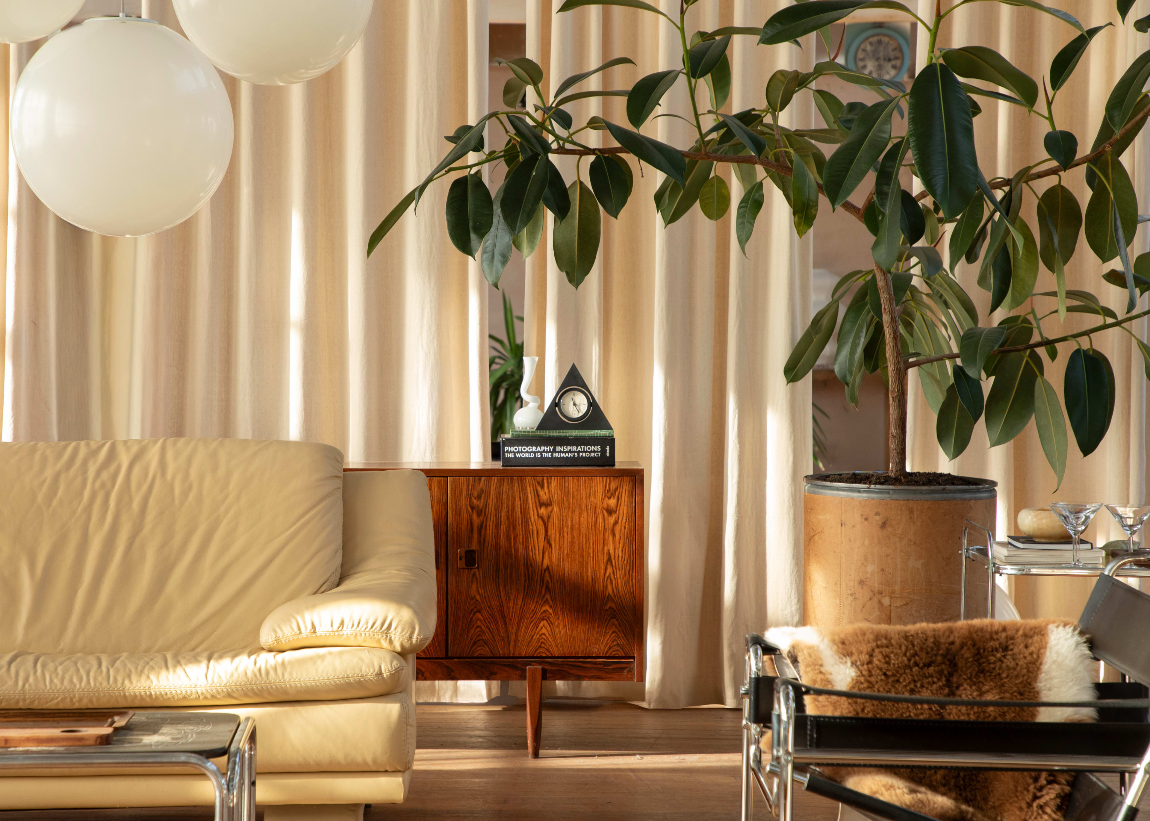 The image shows a living room with a cream-colored sofa, a wooden cabinet, a potted plant, and soft curtains.
