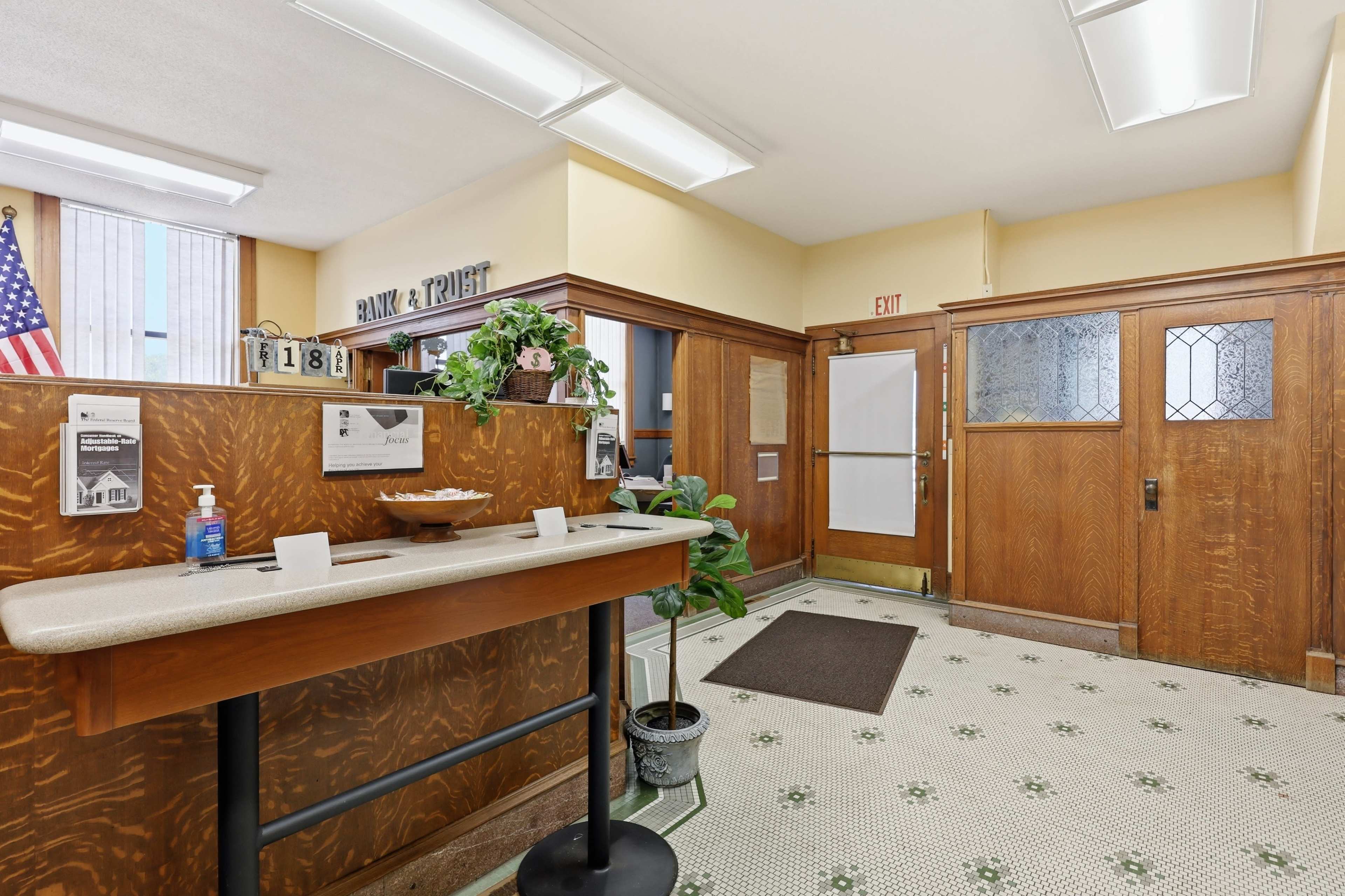 The image shows the interior of a bank lobby featuring a wooden reception desk, a potted plant, and a door leading outside, with decorative tile flooring and a wall-mounted American flag.