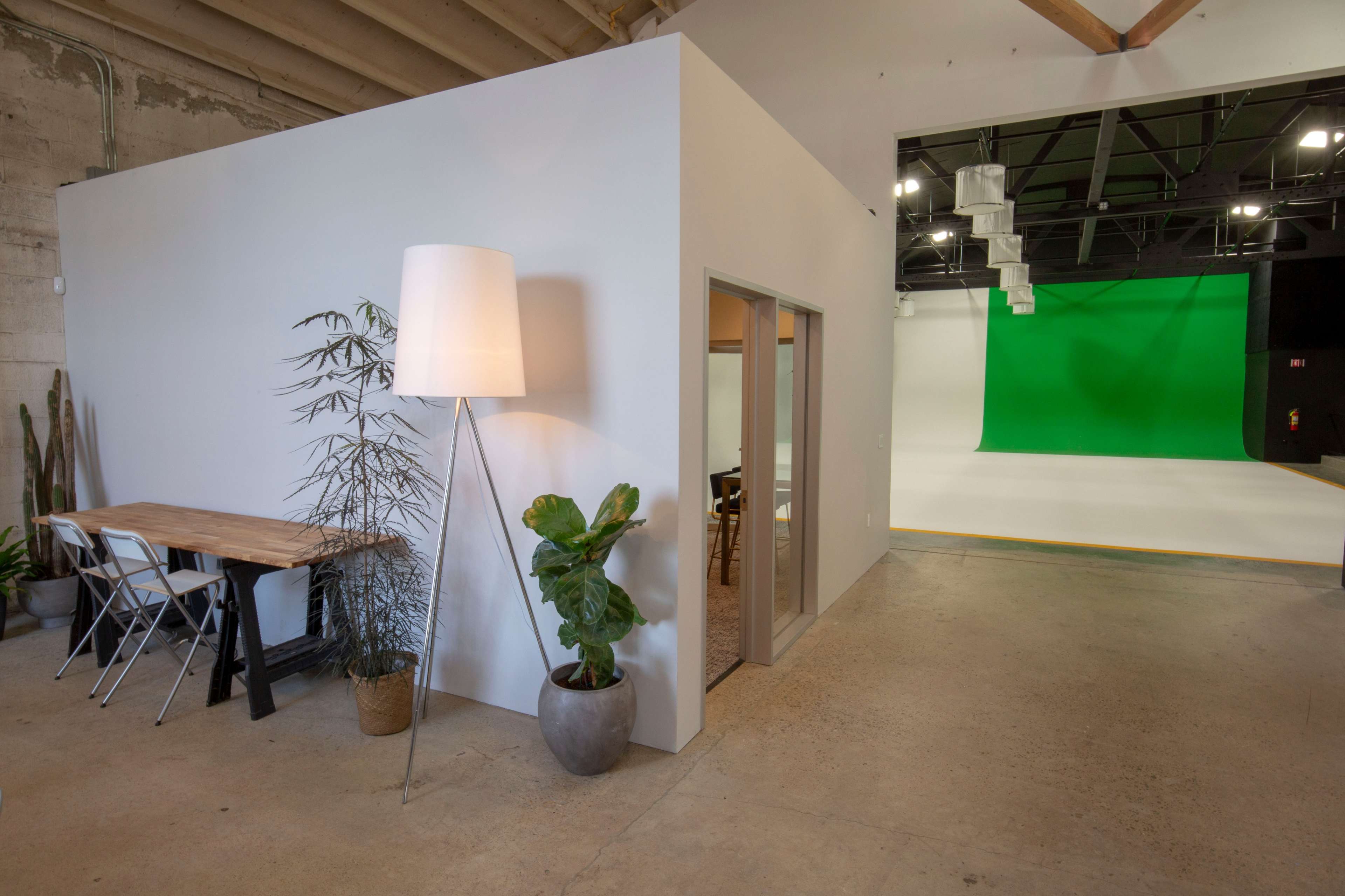 A modern studio space featuring a small white room with a desk and chairs, a tall lamp, a potted plant, and a green screen backdrop.