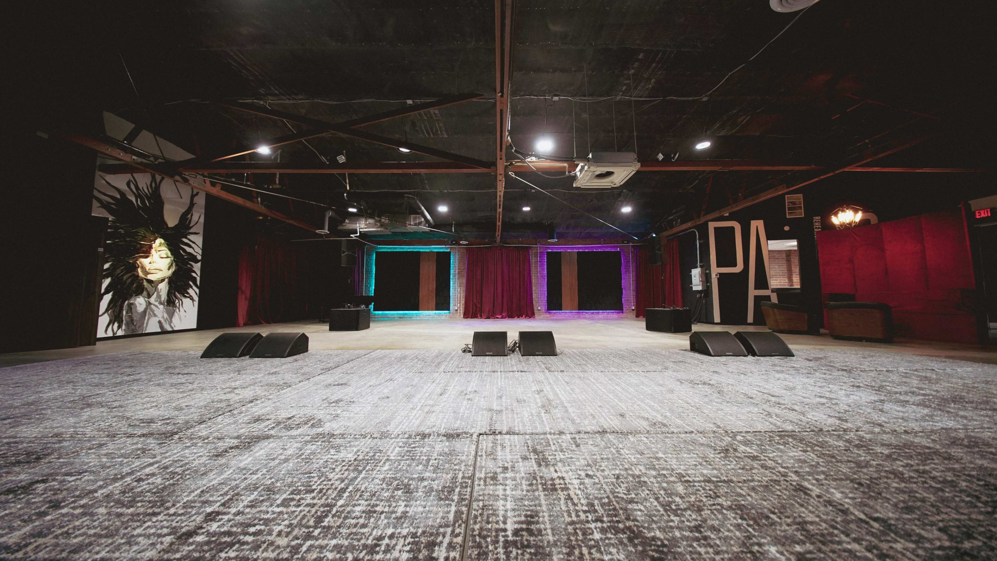 The image shows an empty performance venue with a carpeted floor, a mural on one wall, and stage lighting in the background.