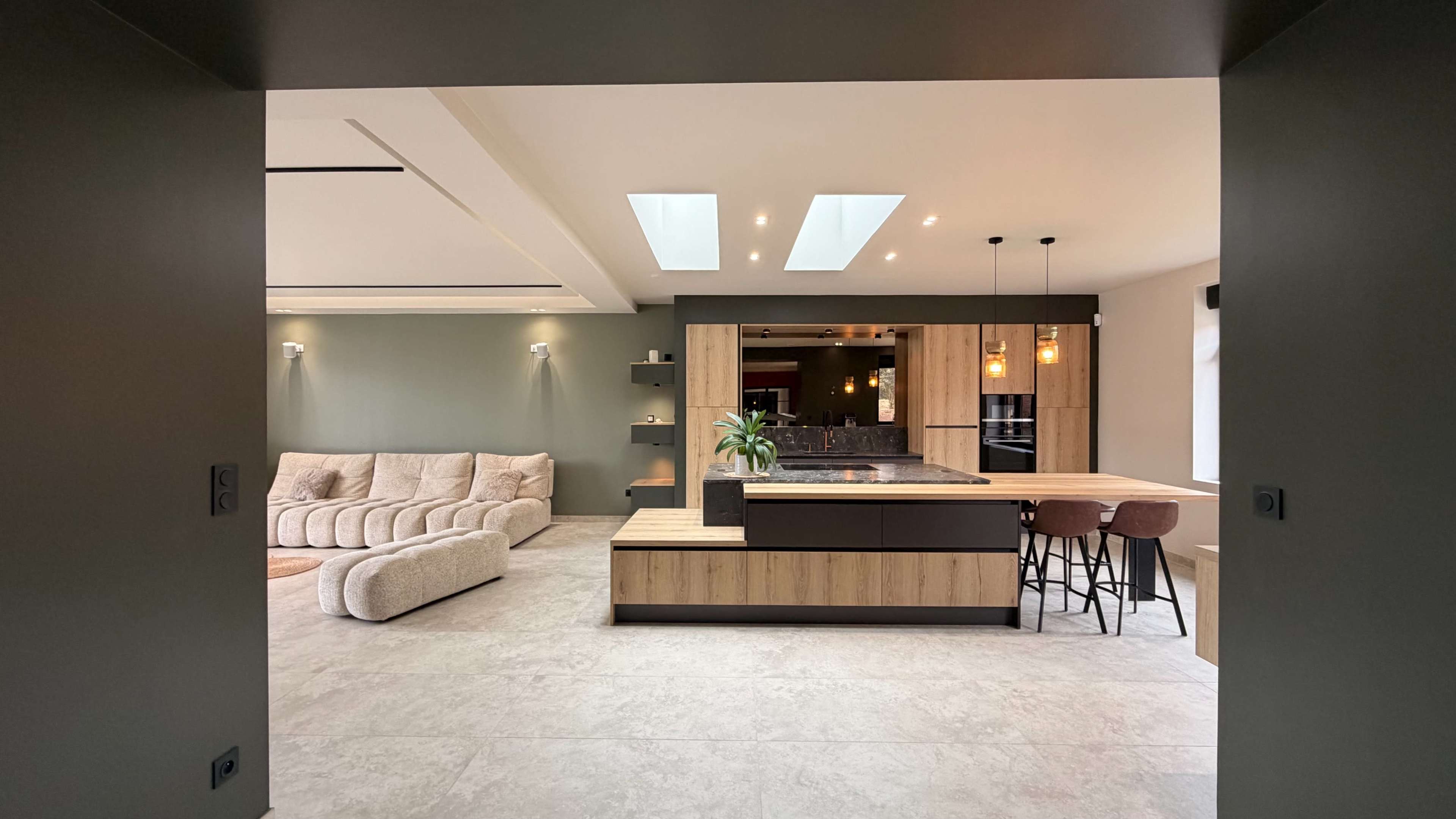 The image shows a modern kitchen and living area with an island bar, dining space, and natural light coming from skylights.