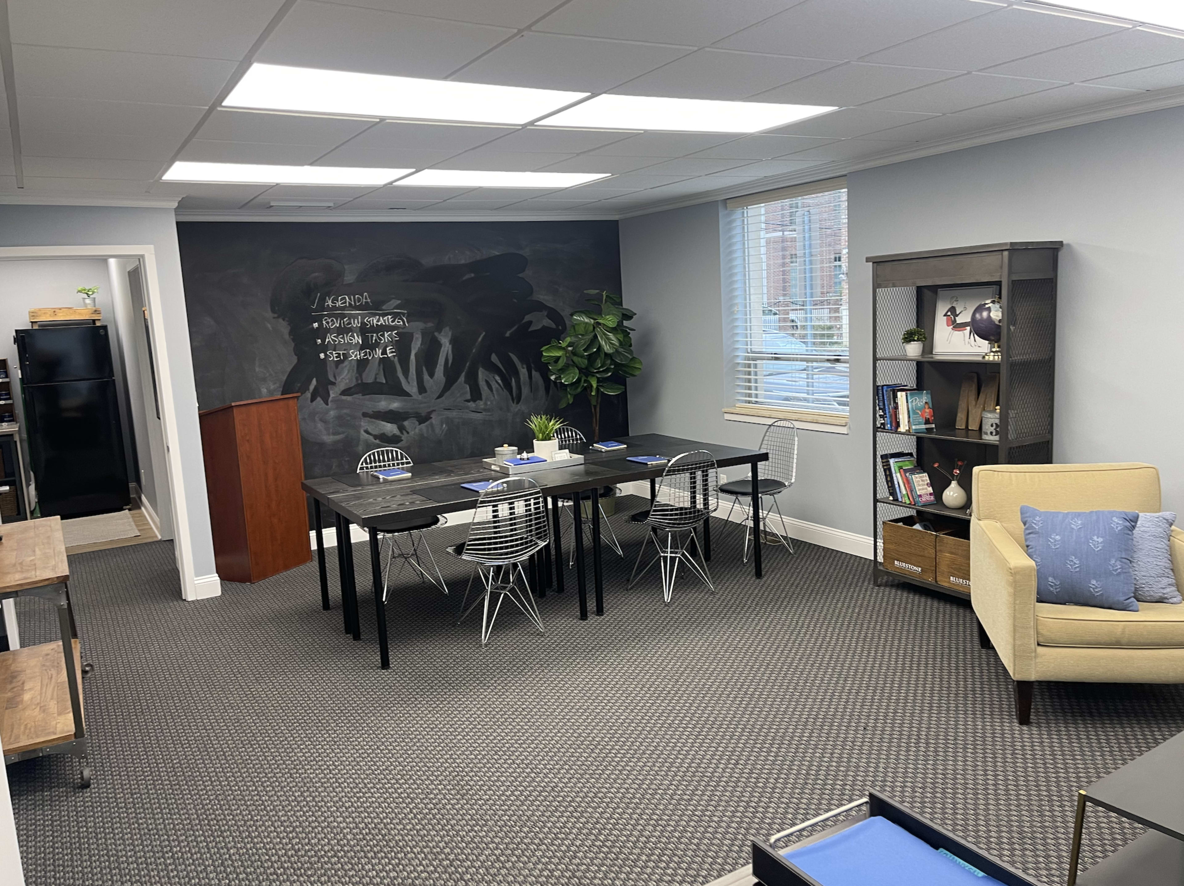 An office space featuring a black chalkboard wall, a dining area with several black tables and white chairs, a small bookshelf, and a beige armchair in one corner.