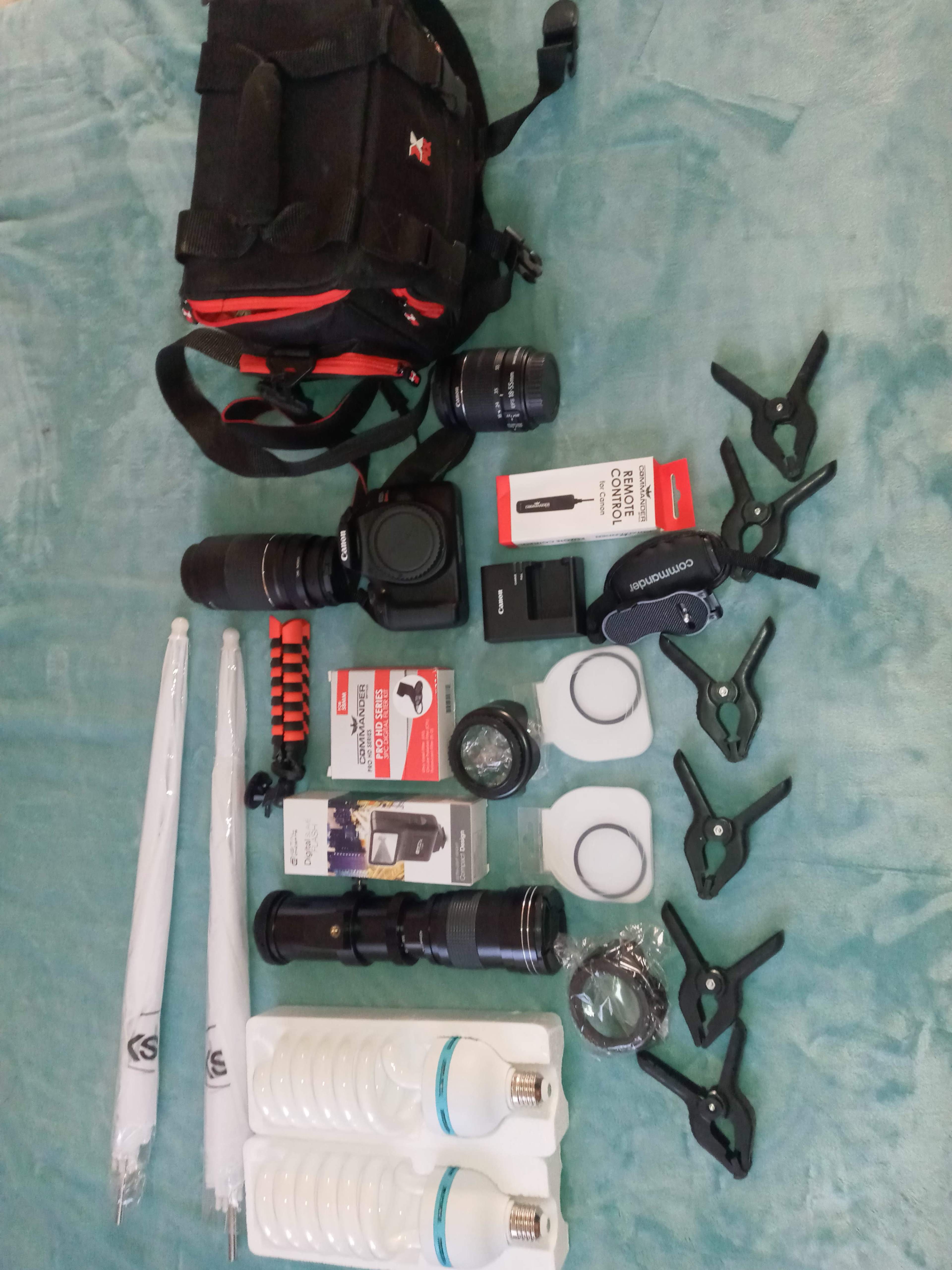 A photography kit with a camera bag, lenses, flashlight, clamps, light bulbs, and various photographic accessories arranged on a light blue cloth.