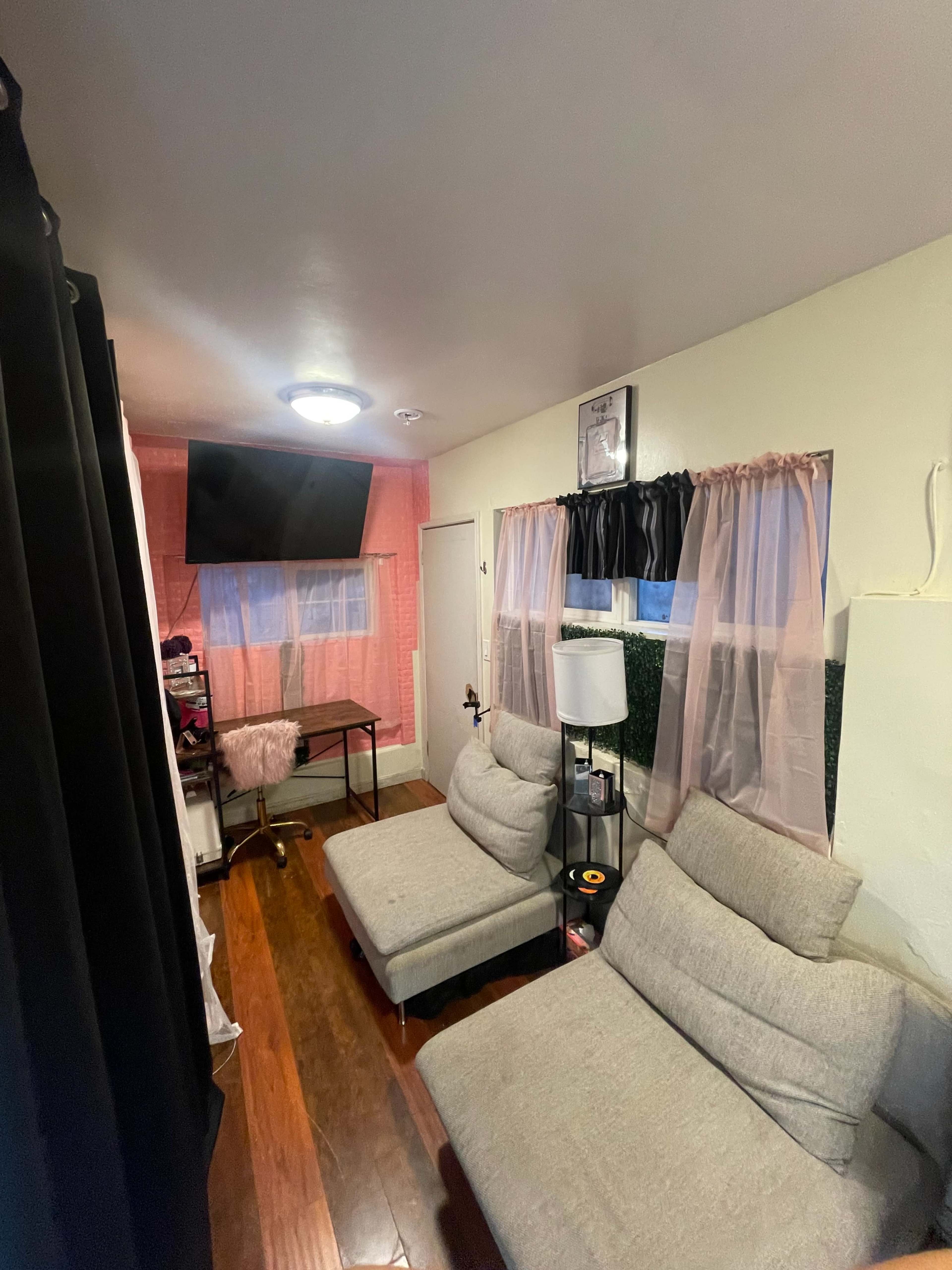 The room features two grey chairs, a flat-screen TV mounted on the wall, and a window with sheer pink curtains.