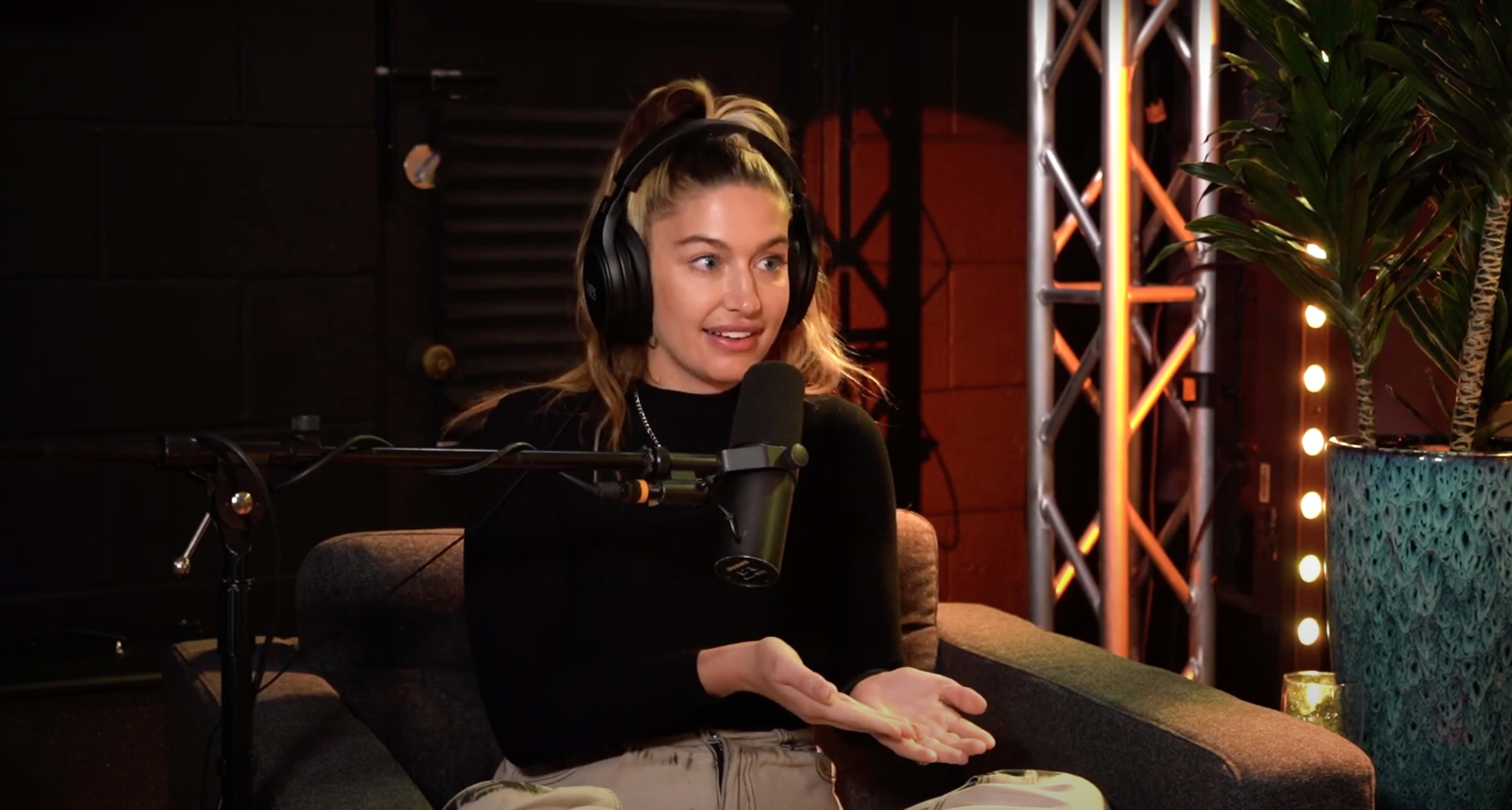 A woman wearing headphones is sitting on a couch, speaking into a microphone during a podcast recording.