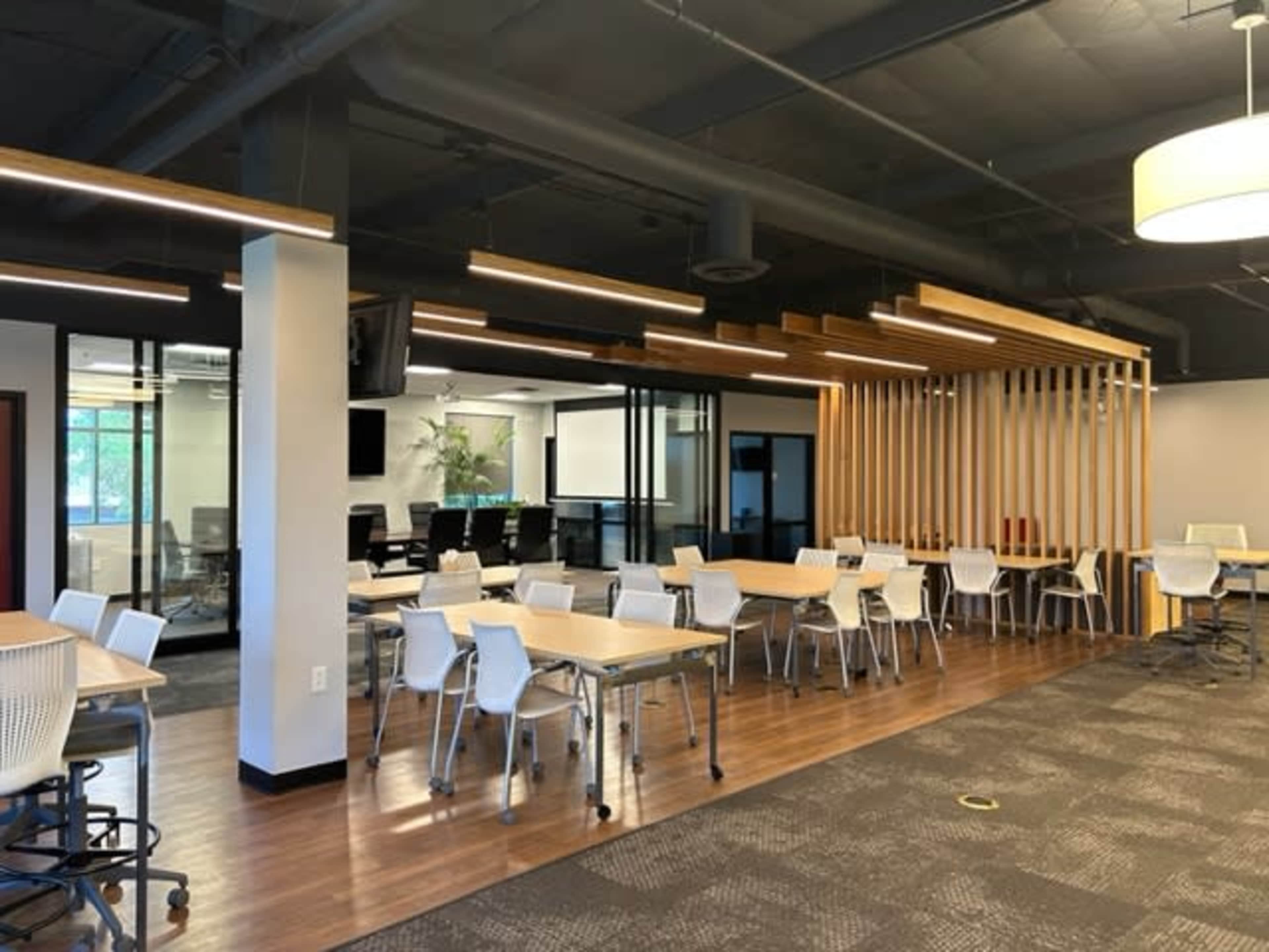A modern office space with multiple tables and chairs arranged in a well-lit area, featuring glass partitions and wooden accents.