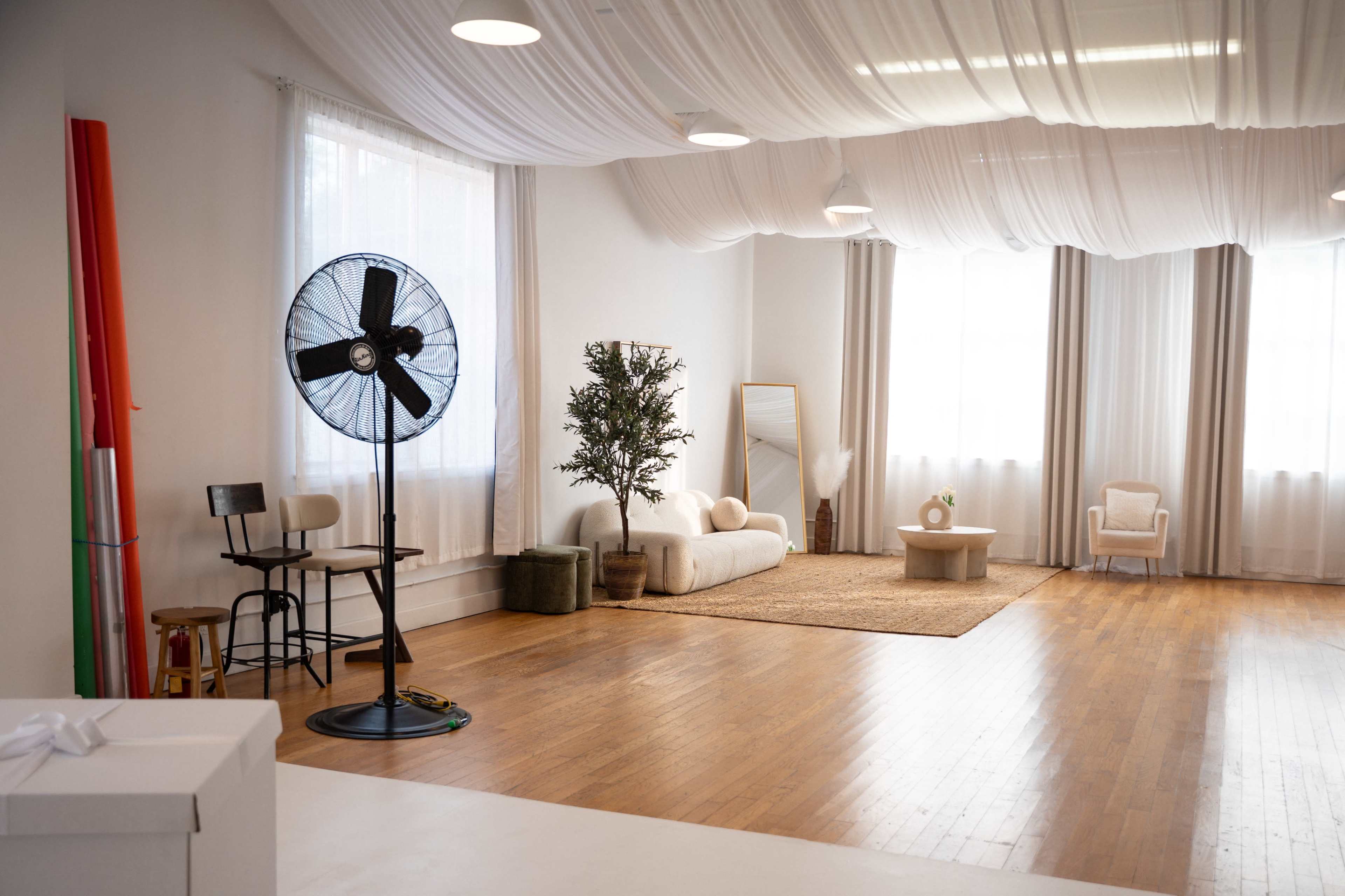 The image shows a bright, minimalist interior space featuring a large fan, a white sofa, a coffee table, and natural light streaming through sheer curtains.