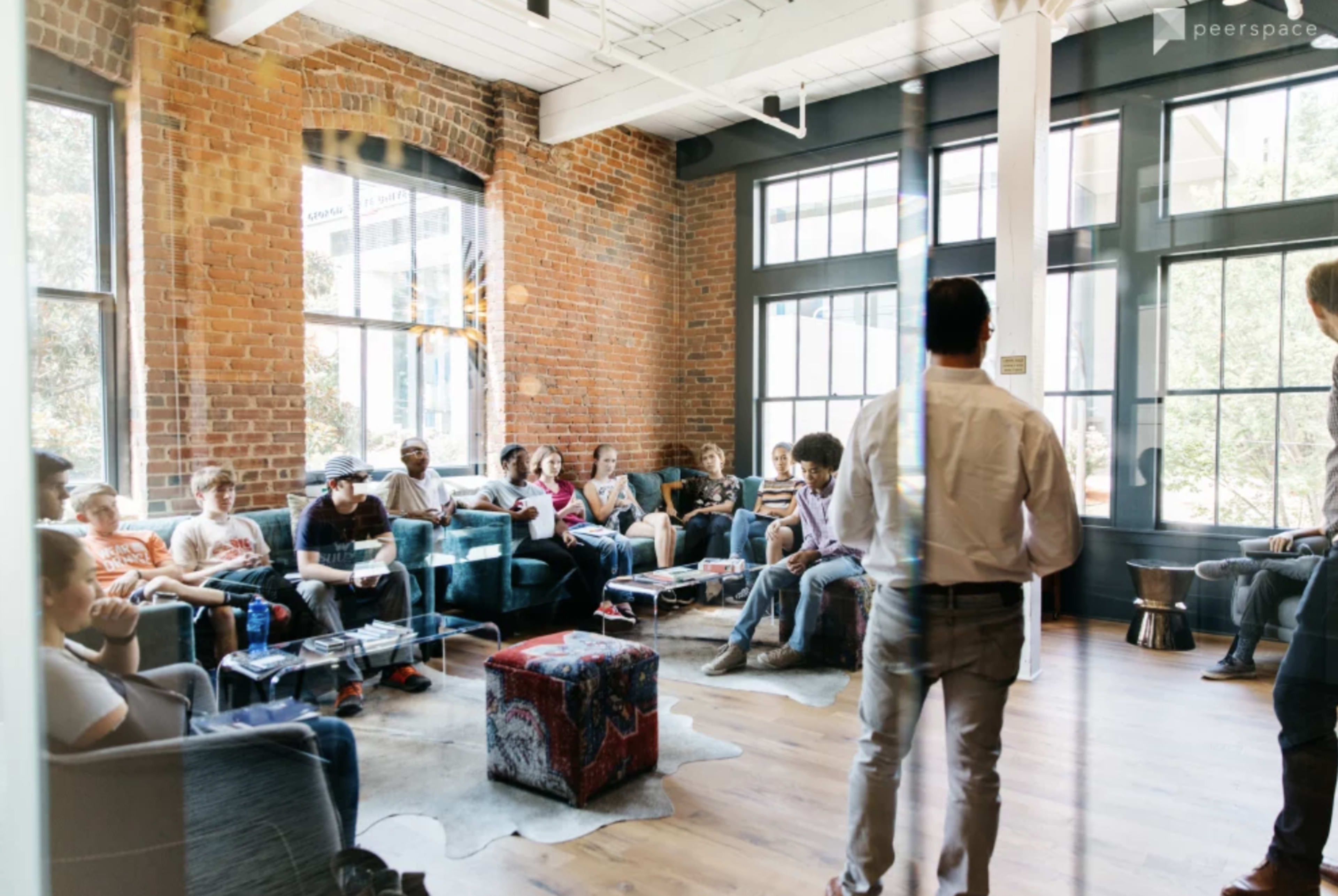 A group of people sits in a bright room with brick walls and large windows, engaged in discussion while a speaker stands in front of them.